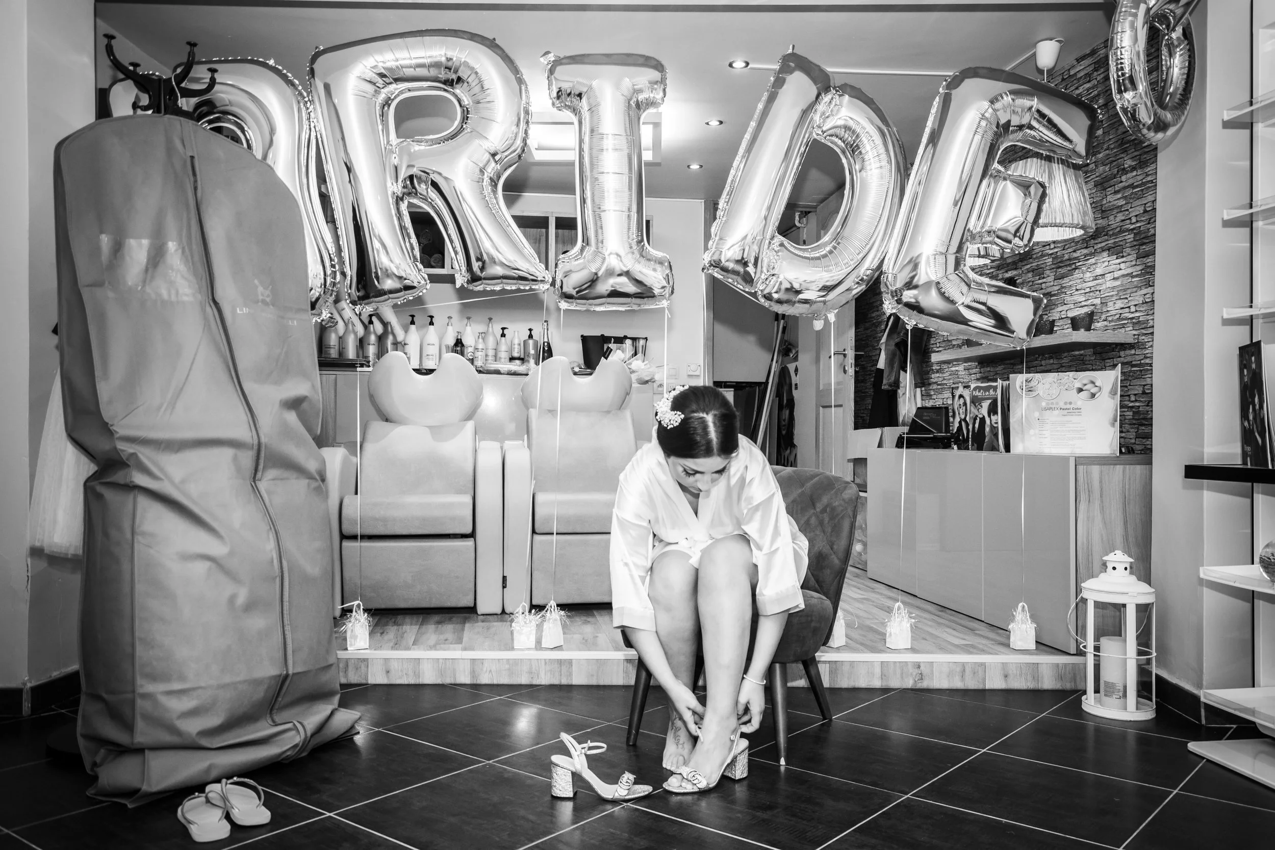 Een vrouw zit op een stoel en doet haar schoenen aan tijdens een feestje. Op de achtergrond zijn grote folieballonnen die samen het woord 'BRIDE' vormen en versieringen, waaronder lampjes en decoraties.