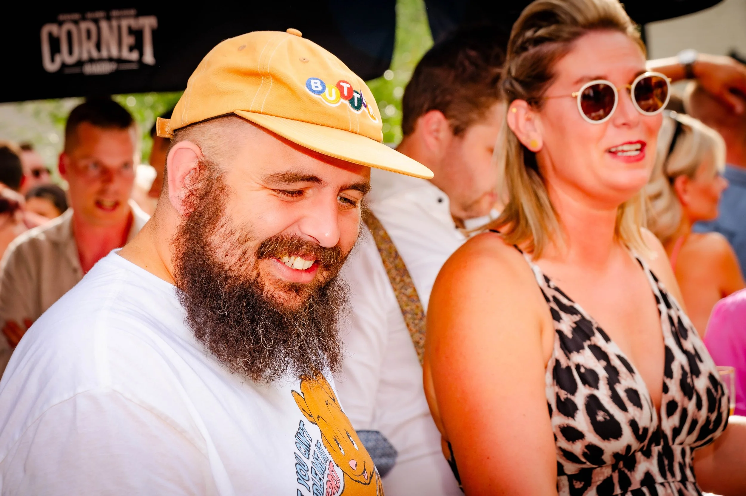 Een groep mensen die genieten van een evenement, waaronder een man met een geel pet en een witte T-shirt en een vrouw met een dierenprint jurk en grote witte zonnebril.