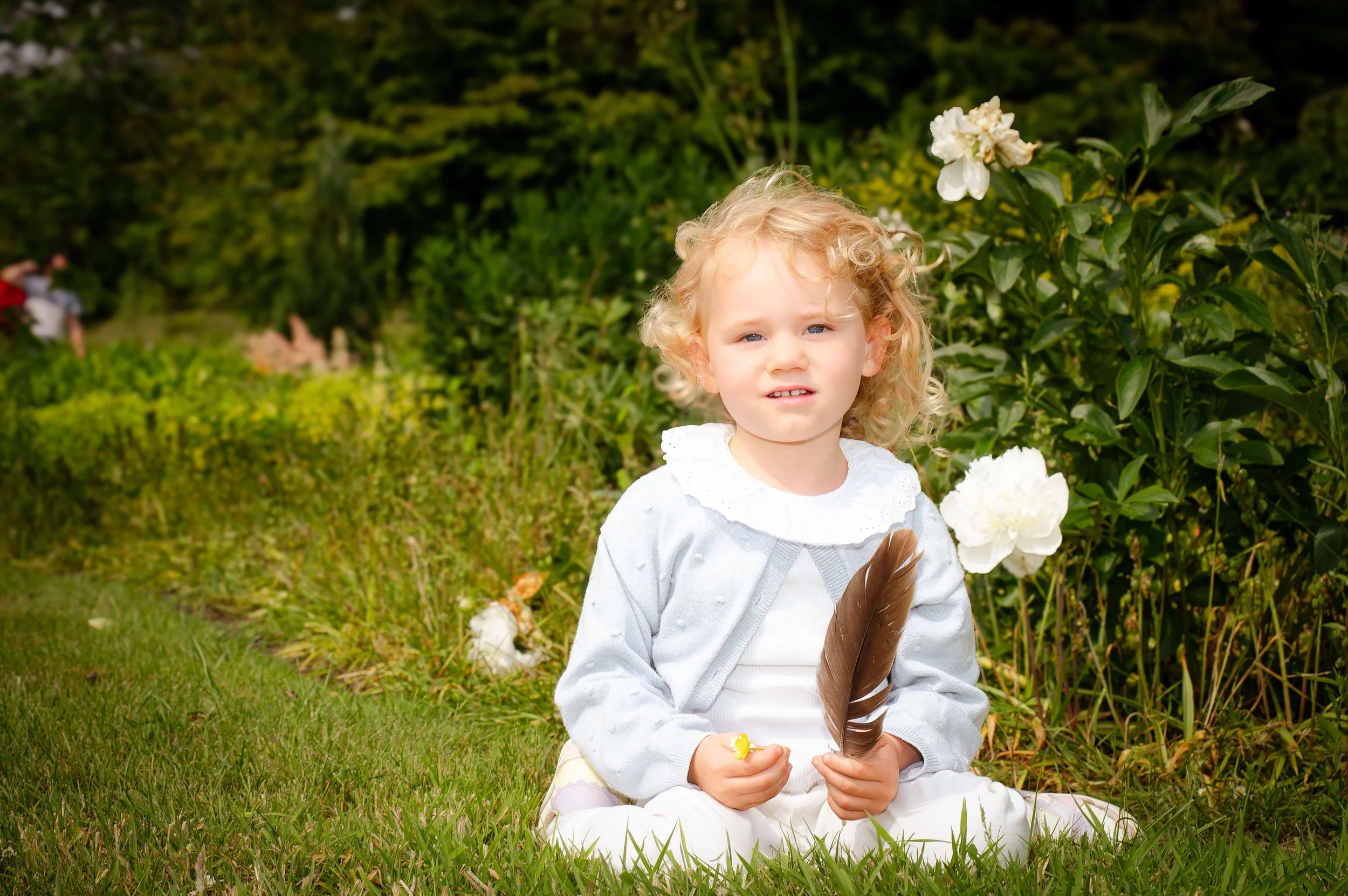 Jongetje met blond haar zit op gras in een tuin, houdt een grote bruine veer en een gele knikker, omringd door witte bloemen, met groene struiken en bomen op de achtergrond.