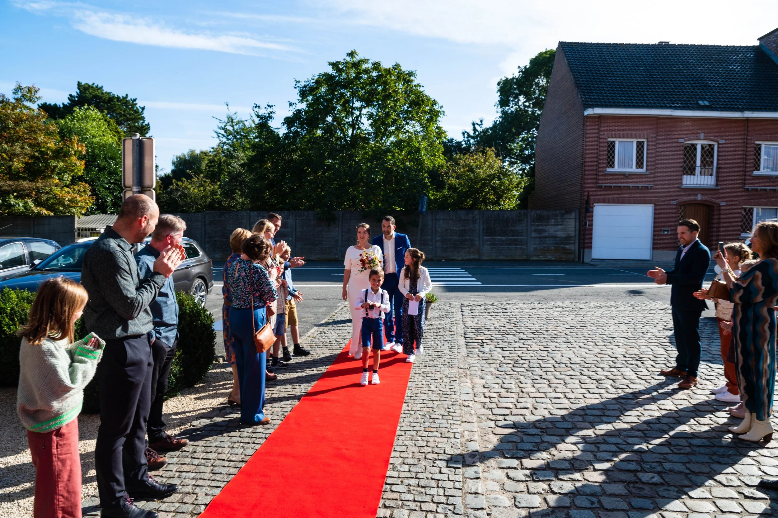 Een bruid en bruidegom lopen over een rode loper terwijl gasten hen aan beide zijden applaudisseren en filmen. Het is een zonnige dag en de gasten staan op een klinkerstraat, met een huis op de achtergrond.