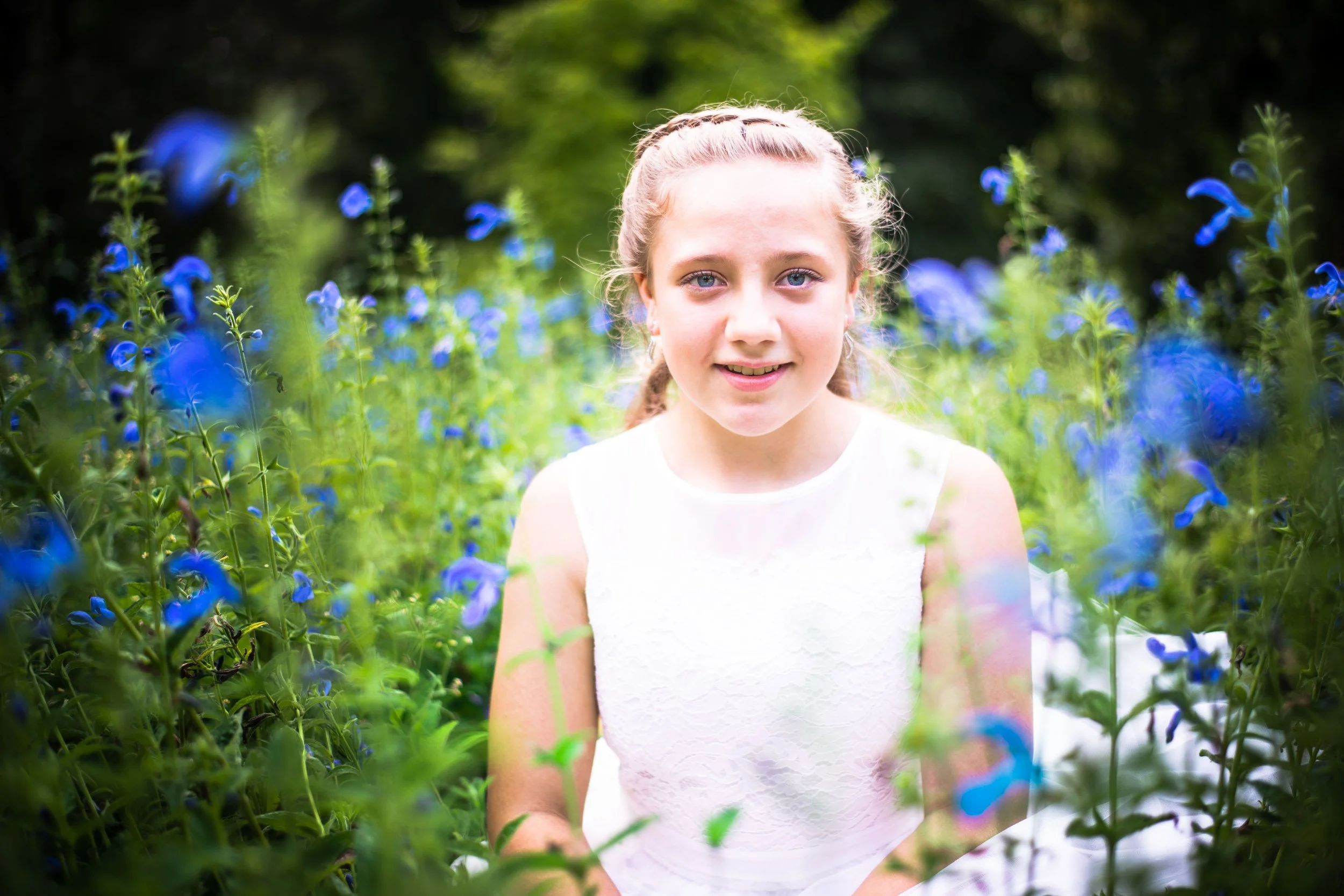 Een jonge vrouw of meisje met lichtbruin haar in een laars, zittend tussen blauwe bloemen in een groene tuin, glimlachend naar de camera.