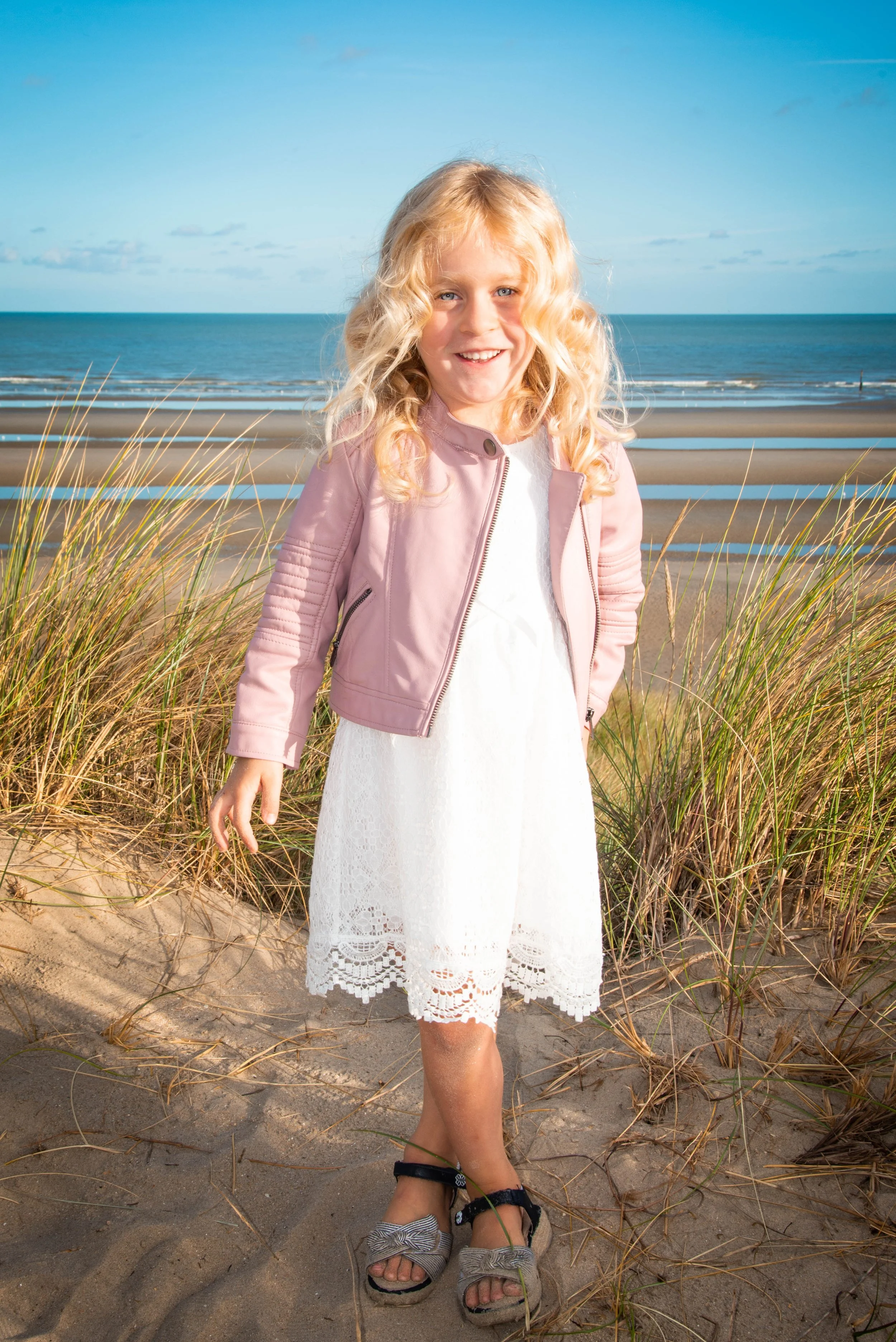 Meisje lachend op het strand met zee en blauwe lucht op de achtergrond, zittend tussen duinen en gras.