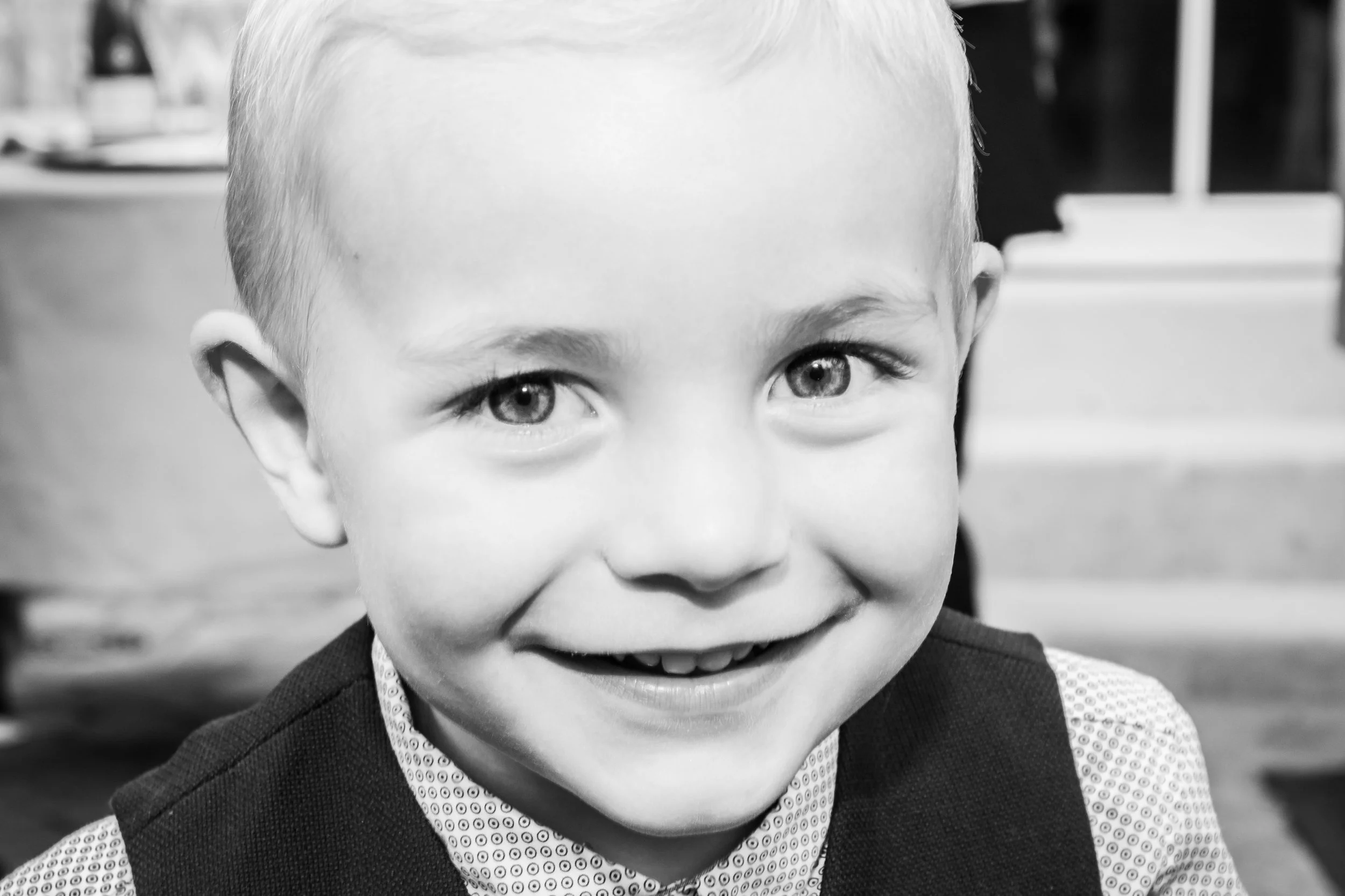 Close-up van een lachende jonge jongen met lichte haar en lichte ogen, gekleed in een overhemd en een bodywarmer, op een binnensetting.