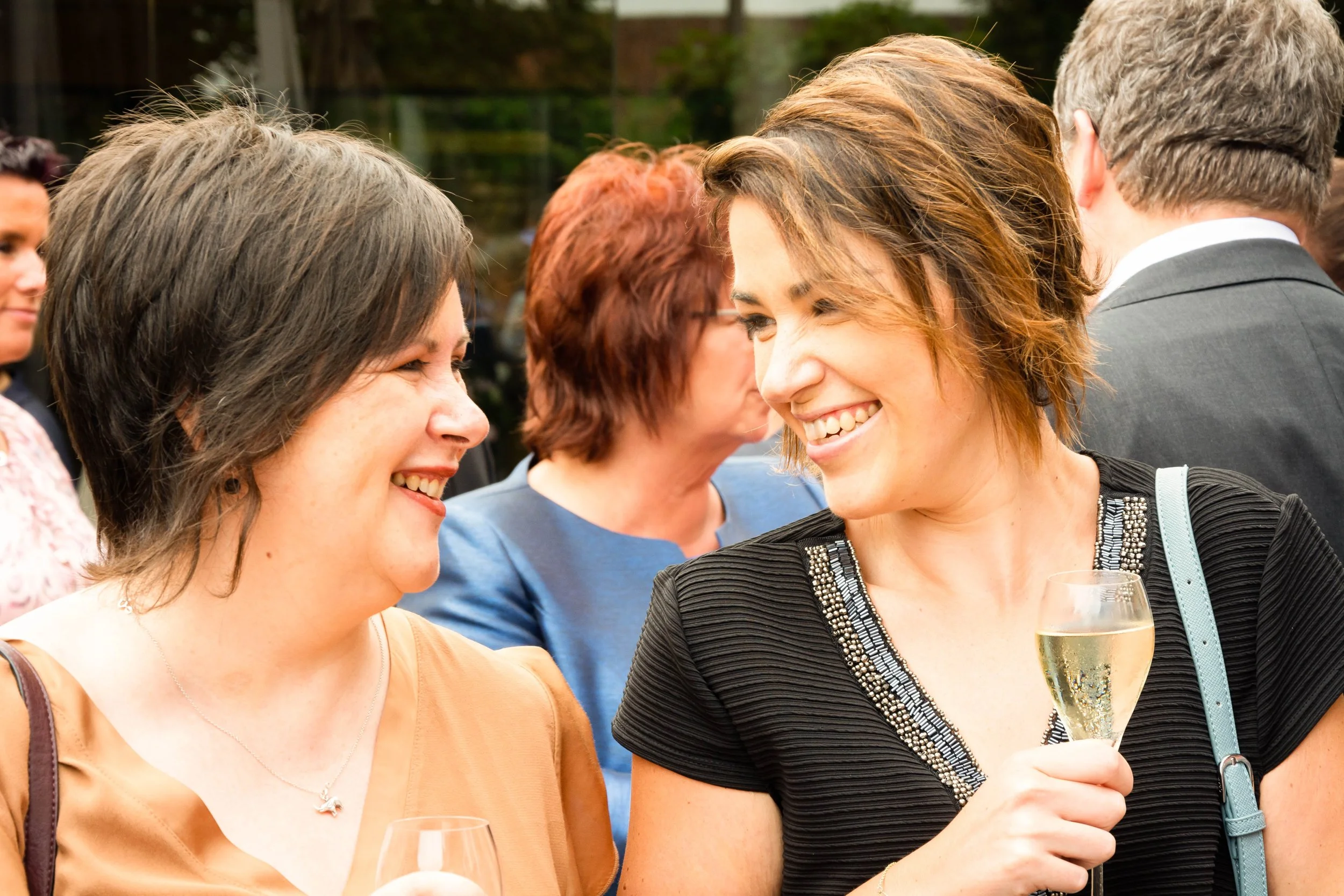 Twee vrouwen lachen en praten tijdens een sociale bijeenkomst, de ene houdt een glas witte wijn vast.
