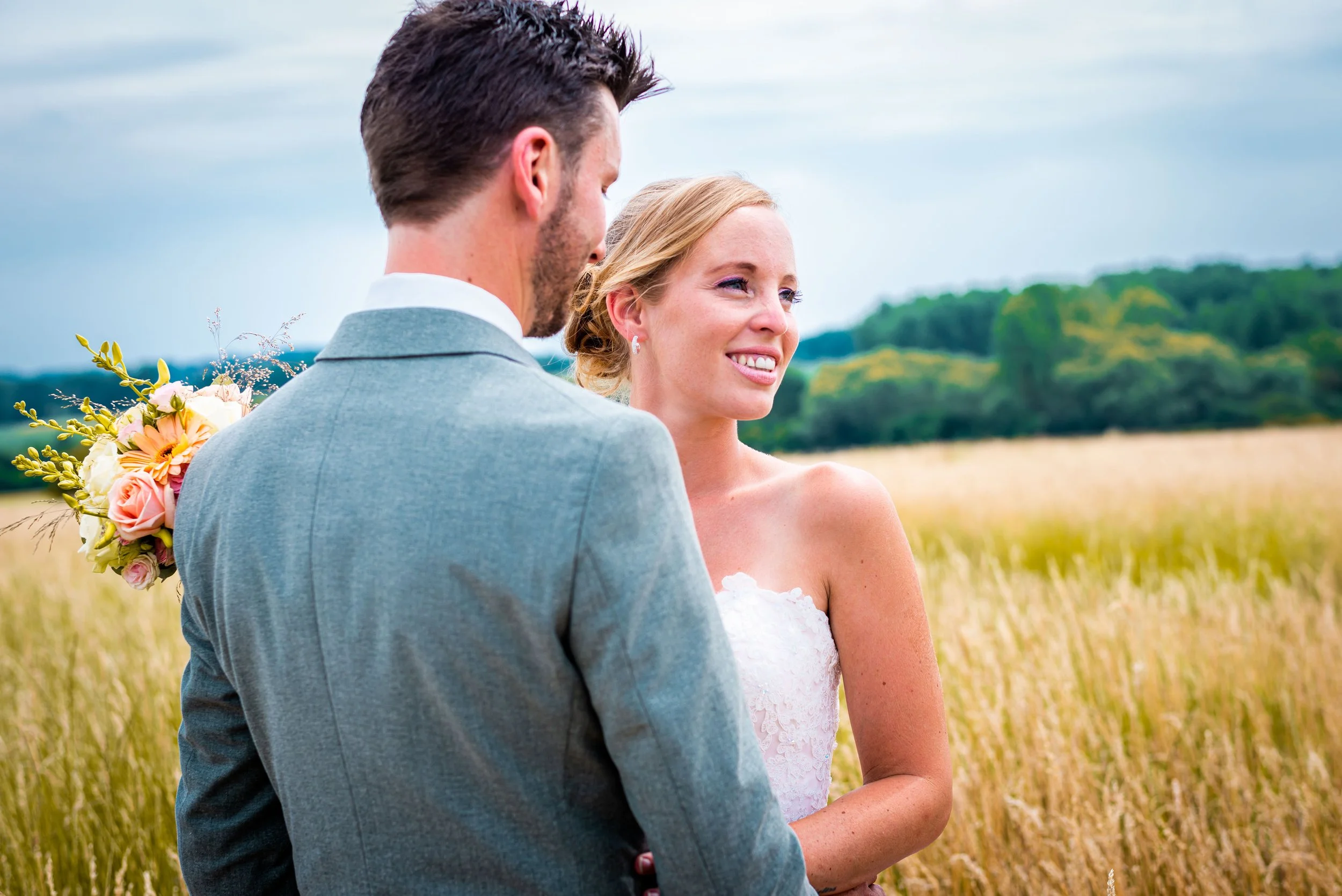 Een stel op hun bruiloft in een veld met goudgele graan, de man in een grijze pak en de vrouw in een witte jurk, beiden glimlachend en in gesprekken.