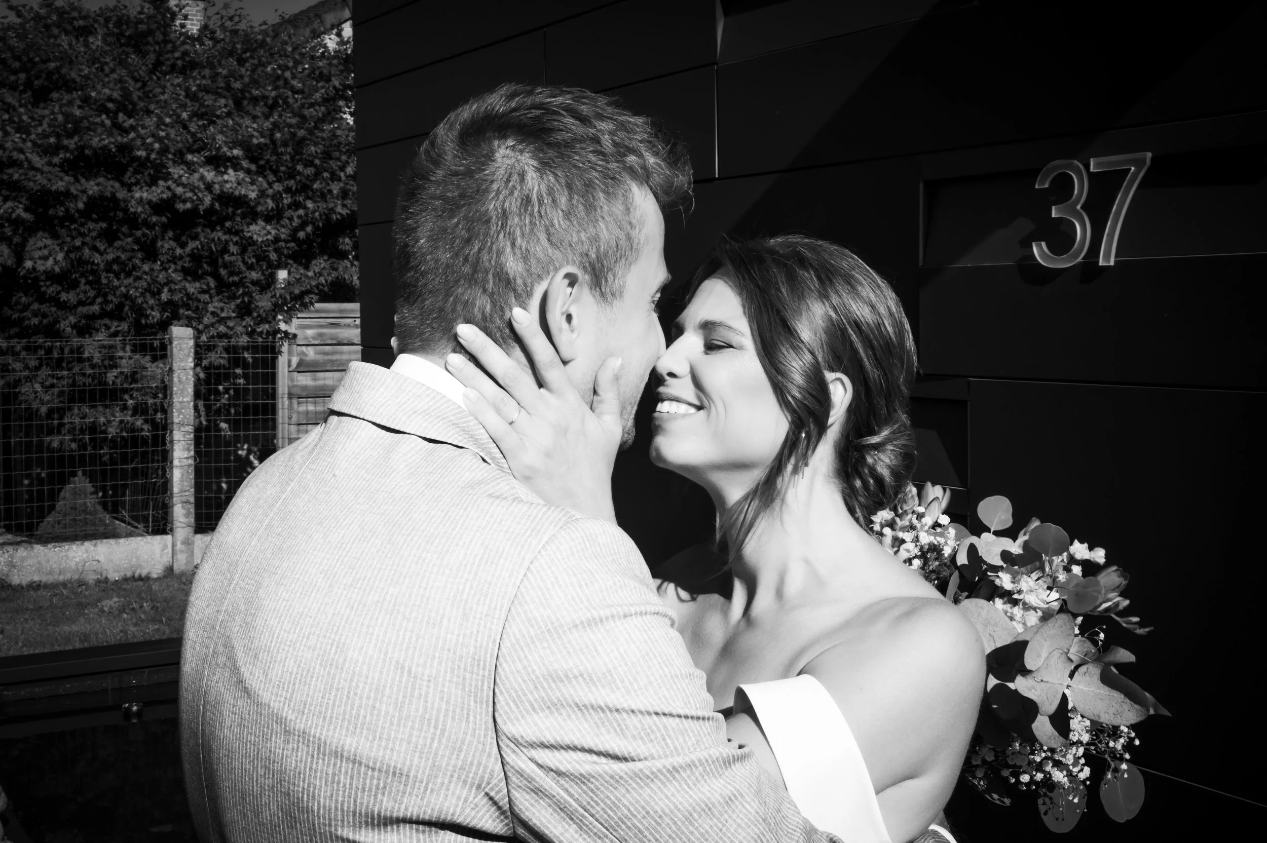 Een zwart-wit foto van een stel die dicht bij elkaar staan, lachend en knuffelend buiten bij een huis met het nummer 37, met bloemen op de achtergrond.