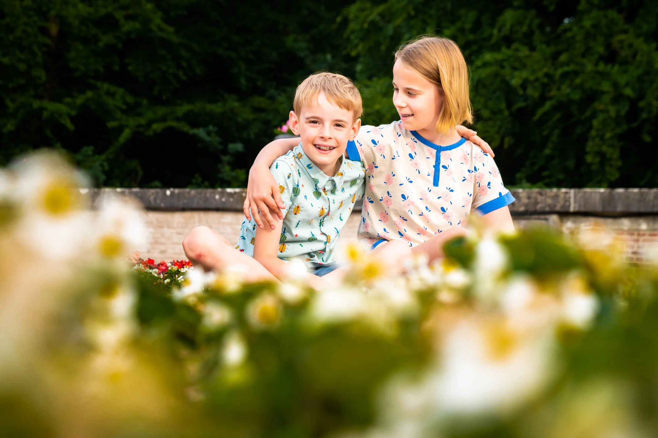Twee kinderen, een jongen en een meisje, zitten samen op een muur met bloemen en een groen bos op de achtergrond. Ze glimlachen en delen een moment van plezier.