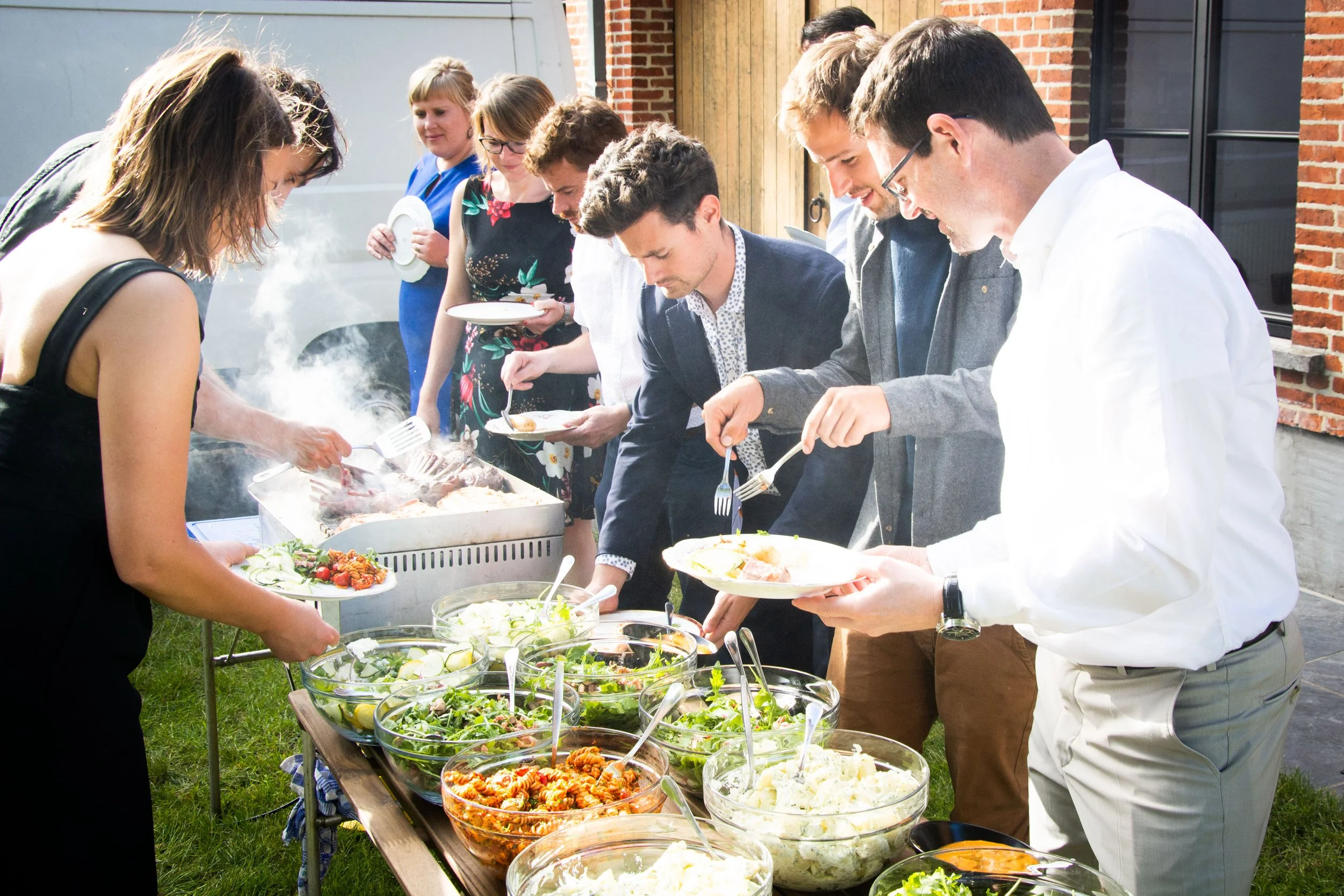 Mensen verzamelen zich voor een buffet met salads en warme gerechten tijdens een barbecuefeest in de tuin.