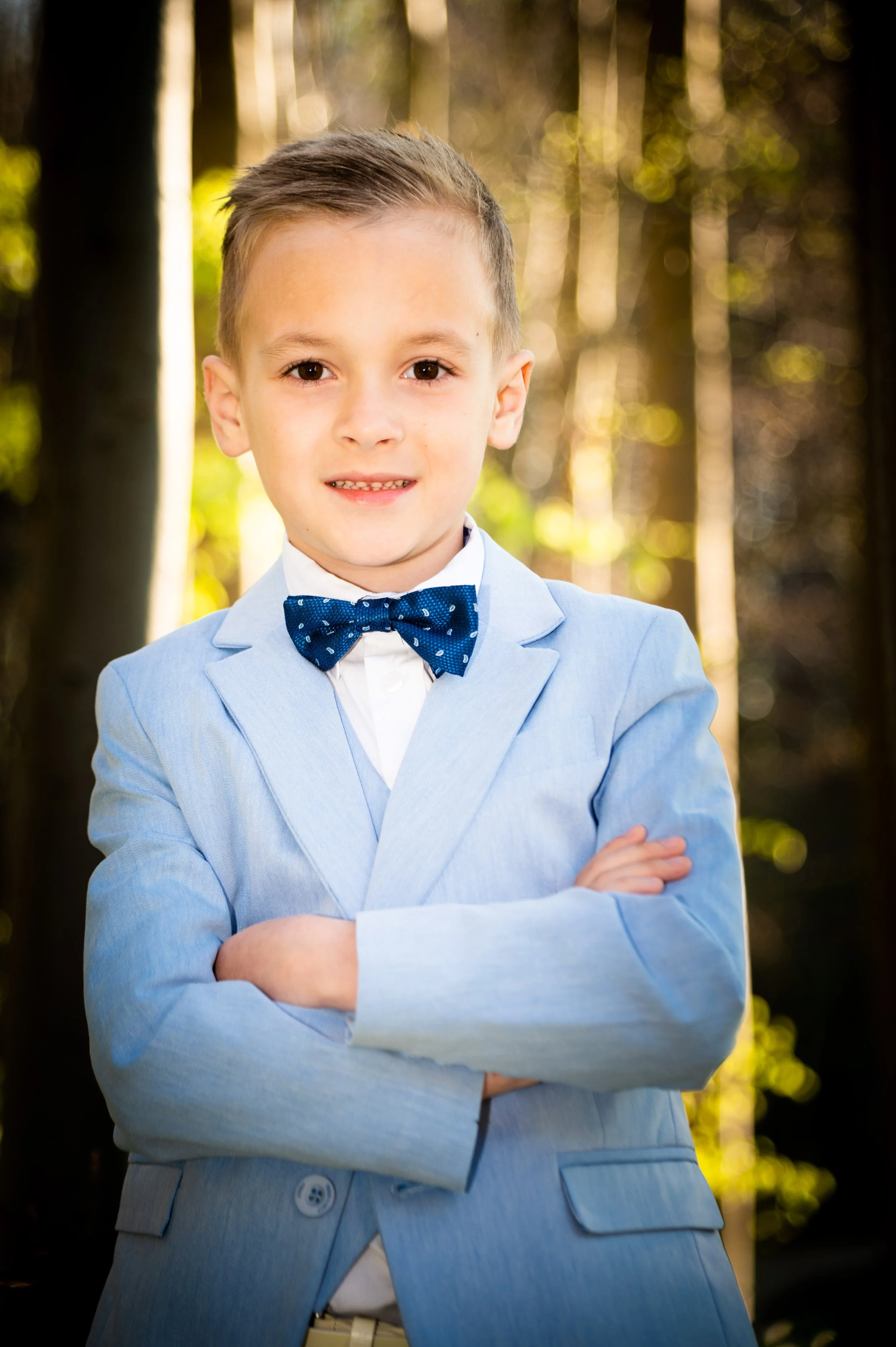 Een jonge jongen in een lichtblauw pak met een blauwe stippen das, staand in een bos met bomen en groen licht dat door de bladeren schijnt.