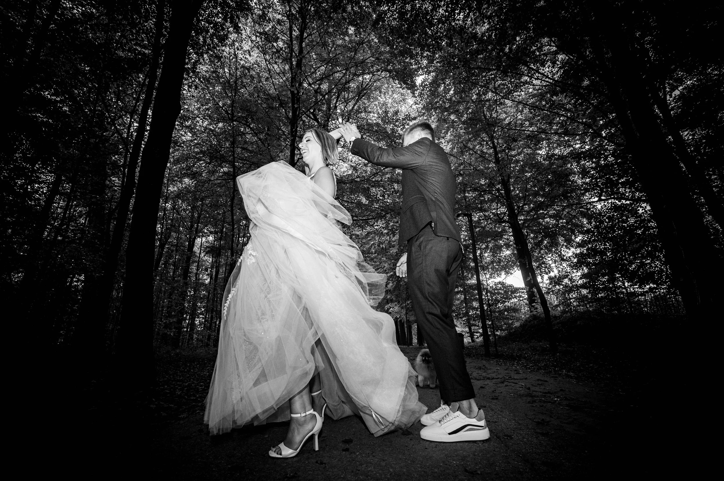 Een getrouwde stel dat dansen in een bos, zwart-wit foto, de vrouw in een trouwjurk en hoge hakken, de man in een pak en sneakers, met een hond op de achtergrond.