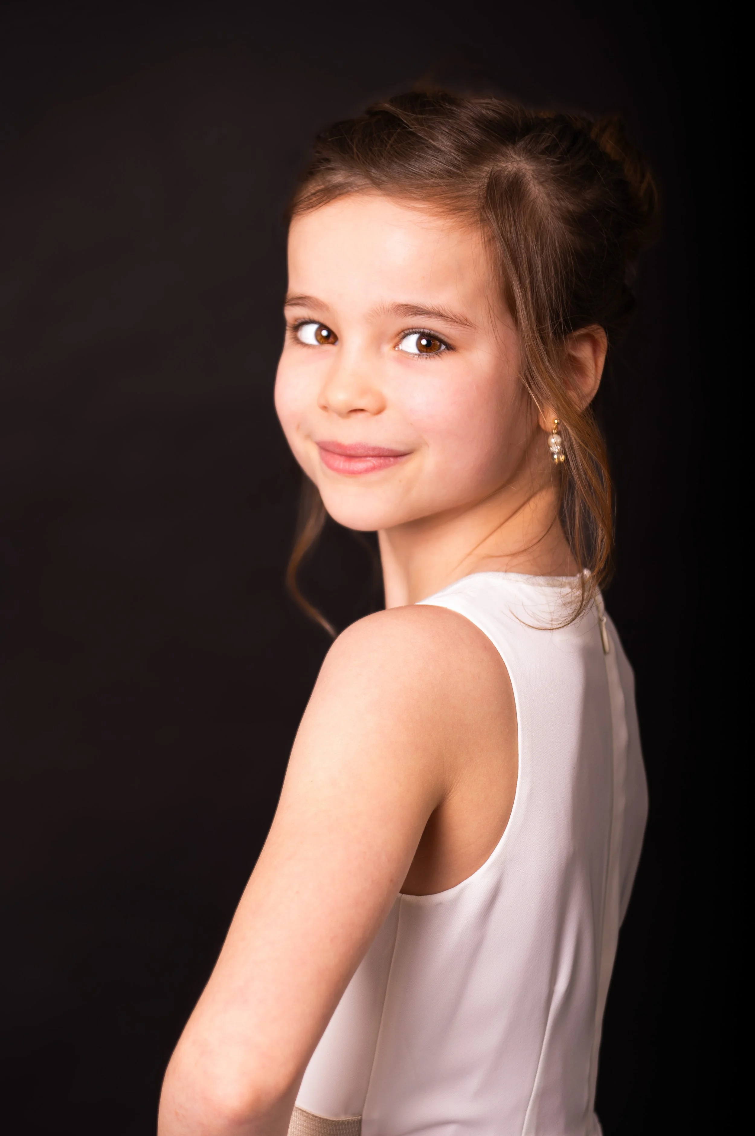 Portret van een jonge meisje met bruin haar en een witte top, lachend tegen een donkere achtergrond.