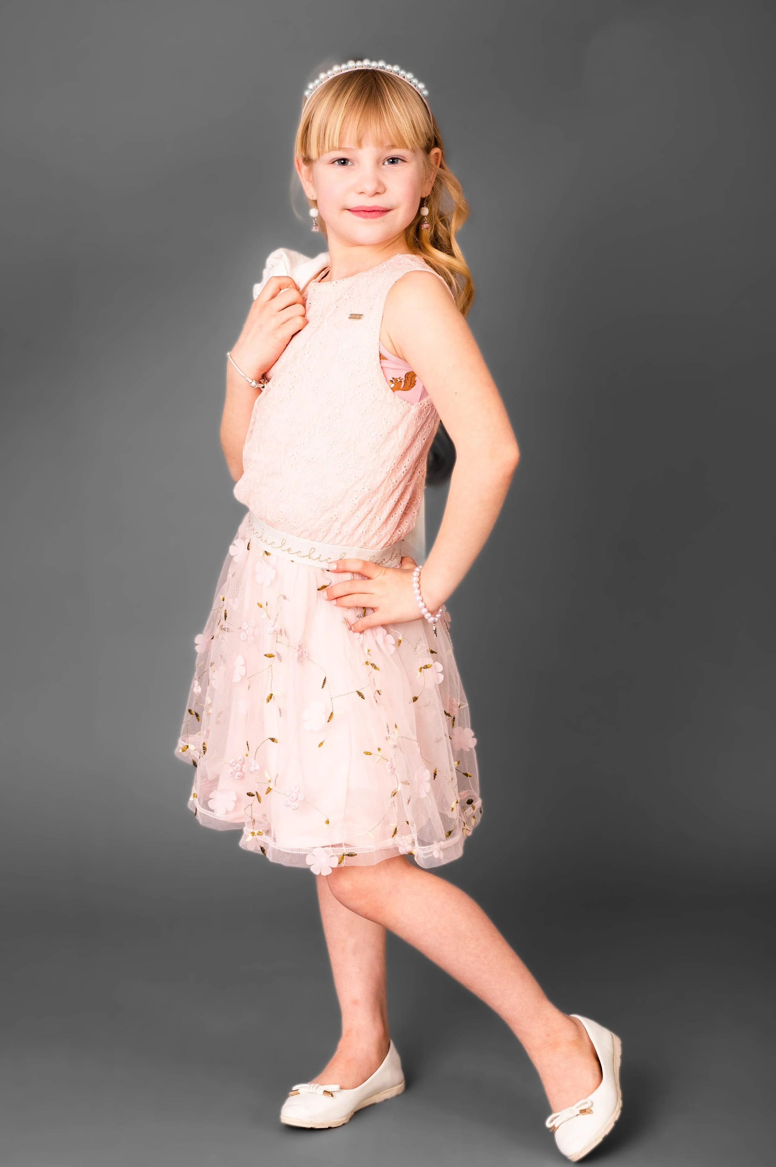 Meisje in zijaanzicht, gekleed in een witte jurk met bloemen, witte schoenen, met een parelketting en tiara, staand op een grijze achtergrond.
