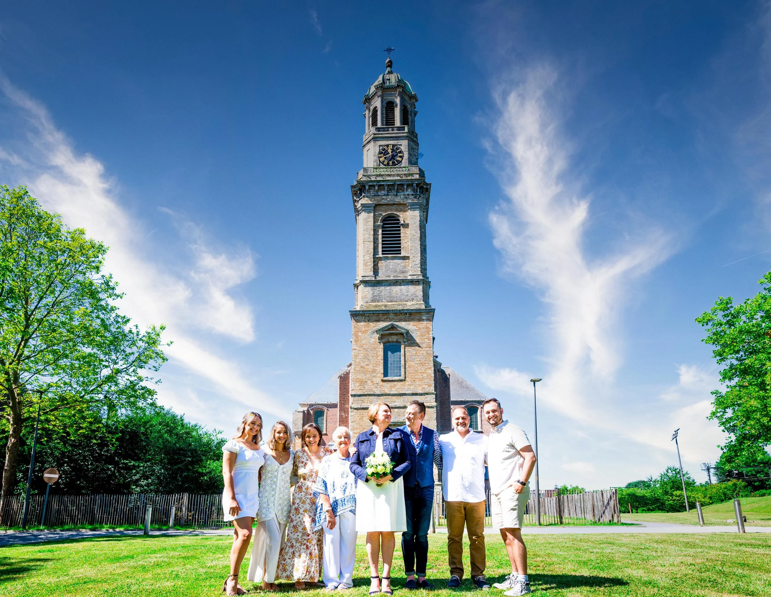 Een groep mensen poseert voor een kerk, met een toren en een blauwe lucht op de achtergrond.