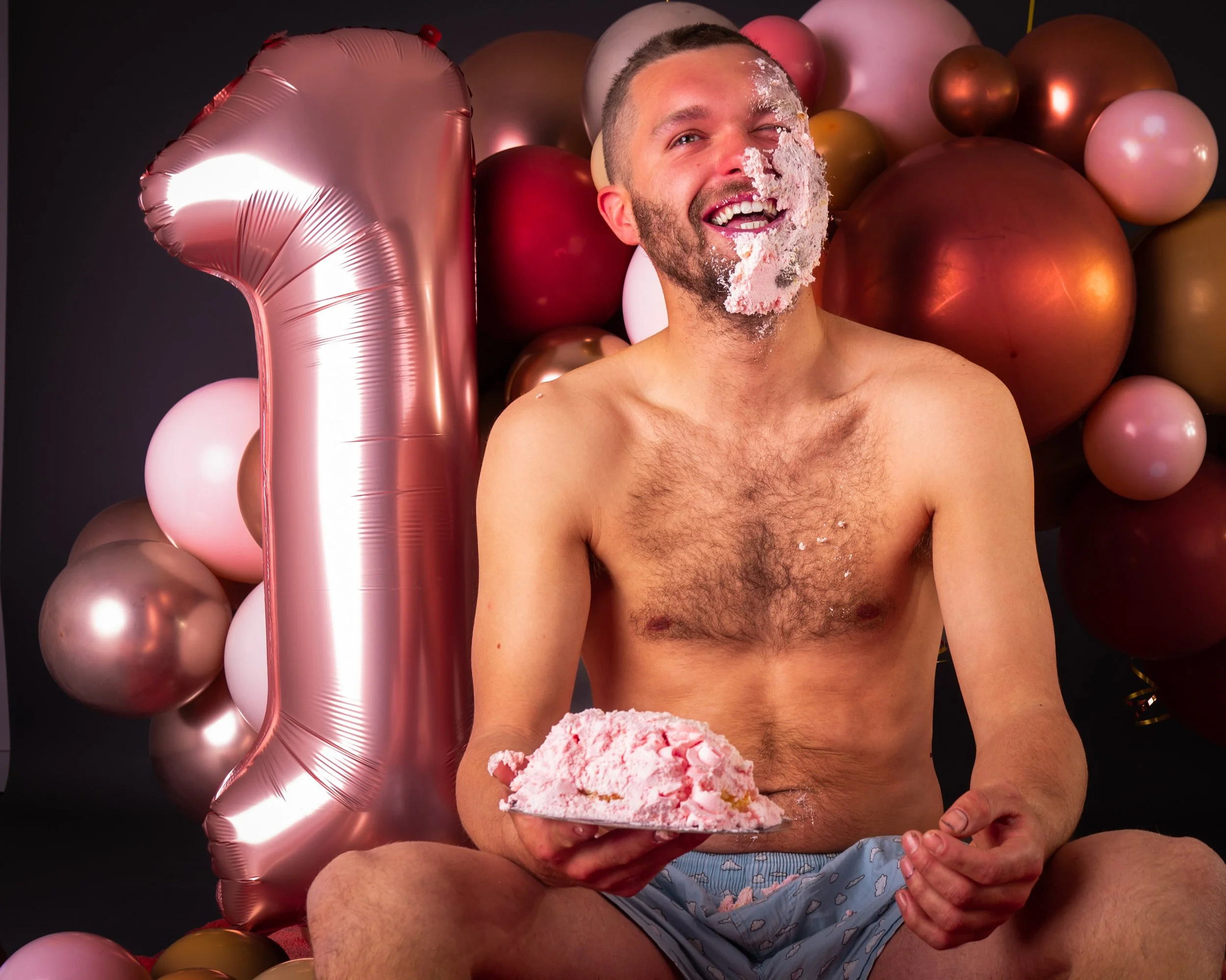 Vrolijke man die op zijn eerste verjaardag viert, zittend met ballonnen en een grote roze '1'-ballon op de achtergrond, terwijl hij cake eet en lacht.