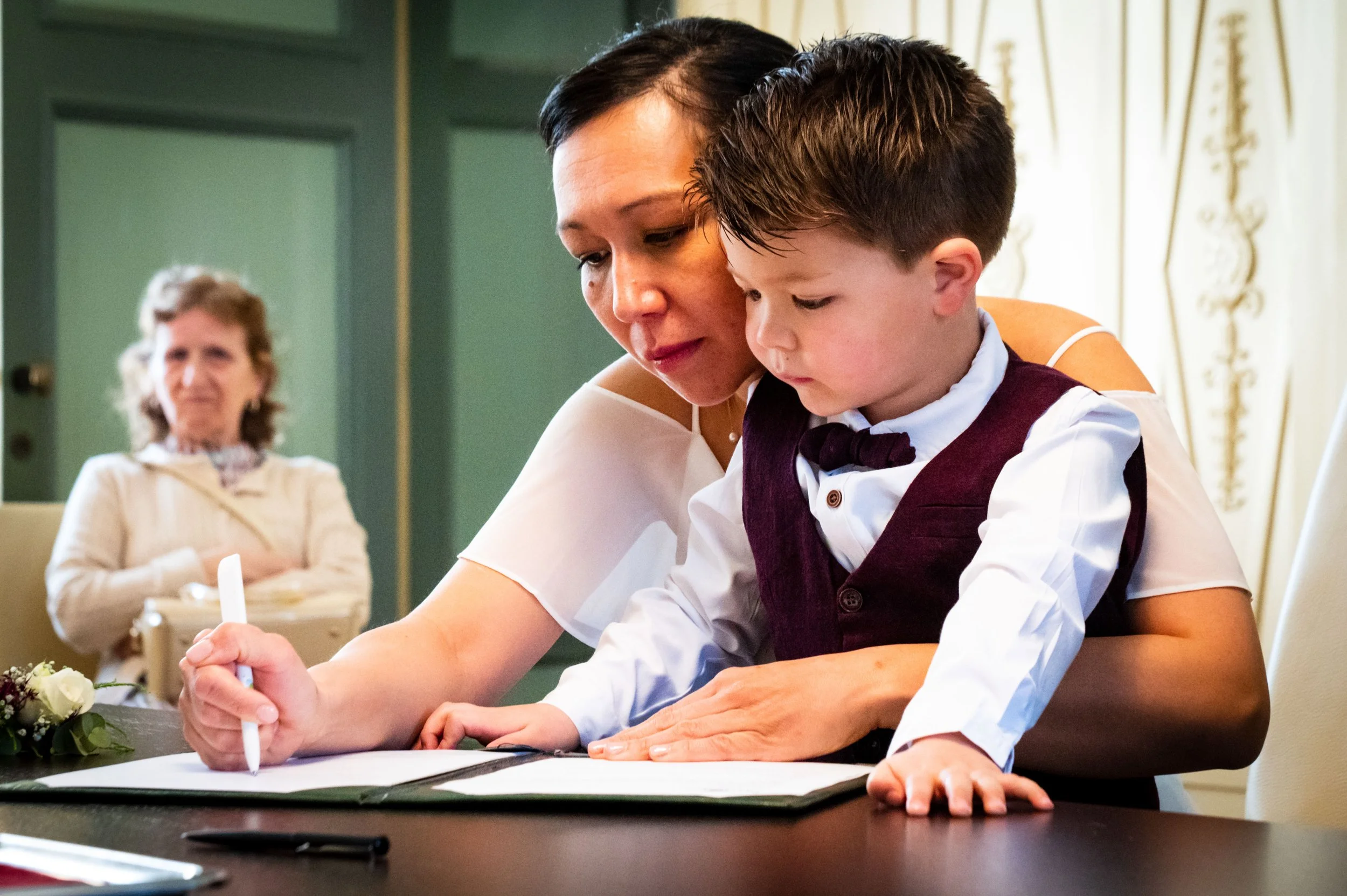 Een vrouw die een jonge jongen helpt bij het ondertekenen van een document tijdens een formele gebeurtenis, met een oudere vrouw kijkend op de achtergrond.