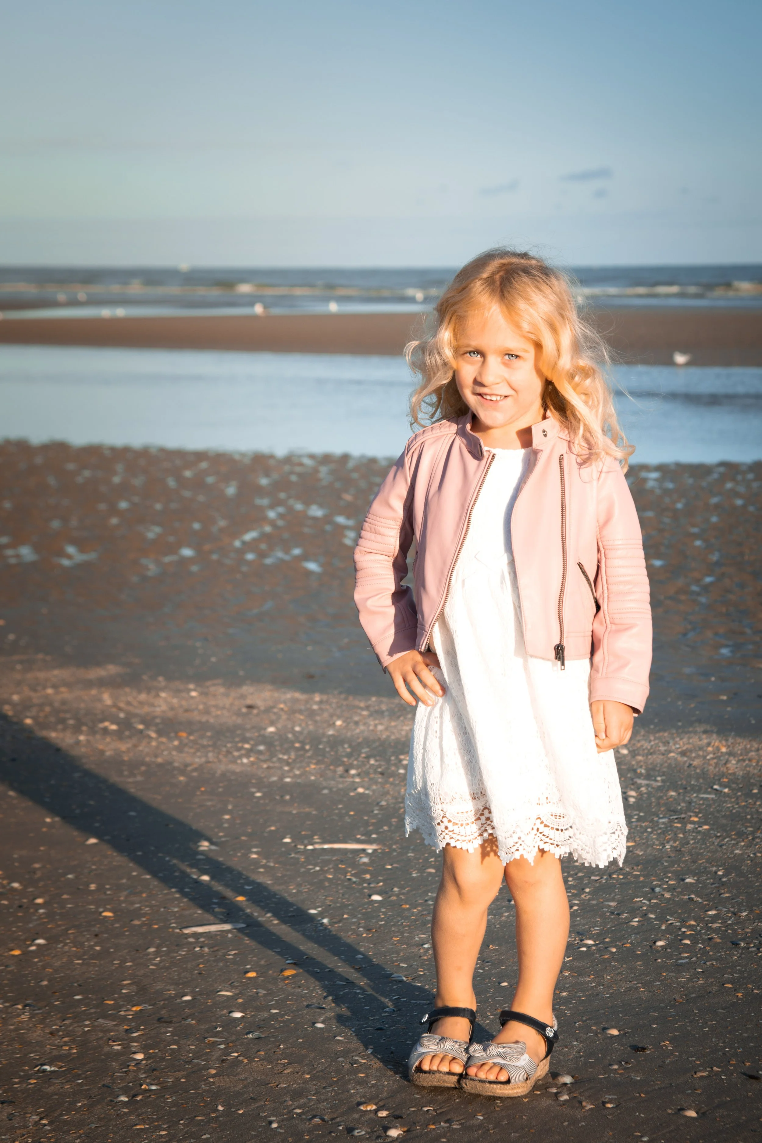 Meisje met blonde krullen, in een witte jurk en een roze jas, staat op het strand naast het water, met een scheve glimlach.