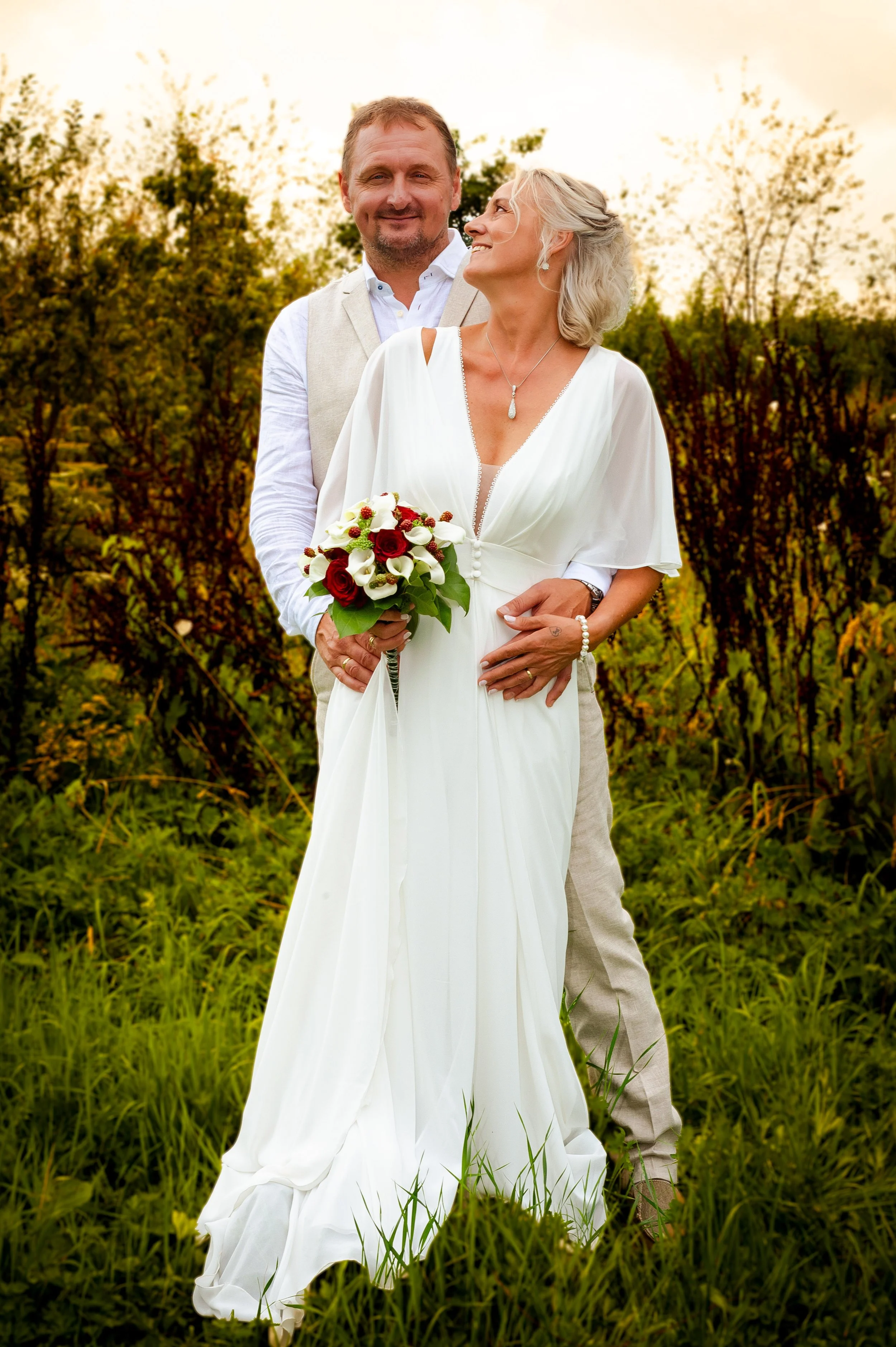 Een oudere man en vrouw in trouwkleding poseren in een grasveld met struiken op de achtergrond, de vrouw houdt een bloemenboeket vast.