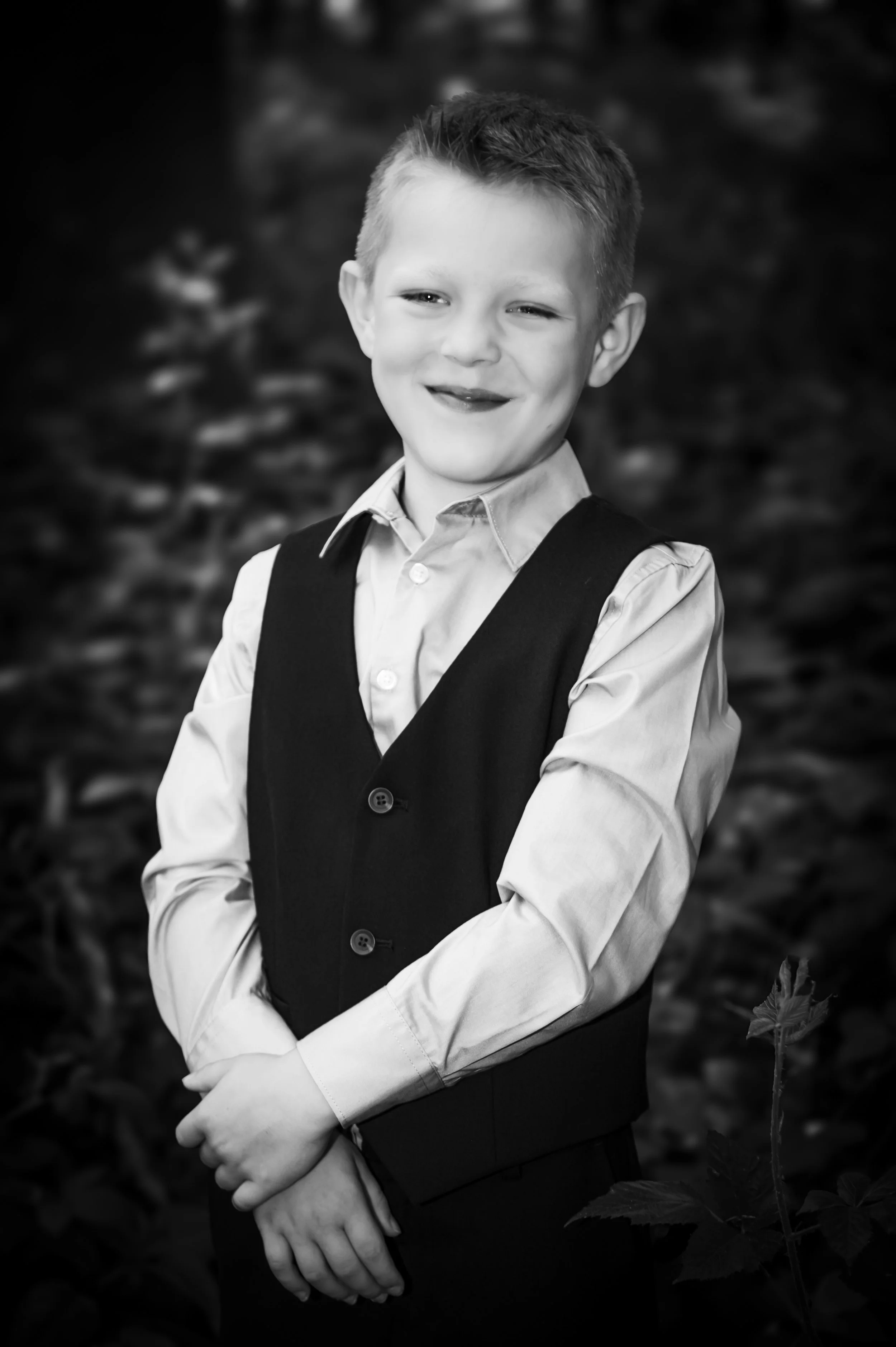 Zoon van een jongen in formele kleding, glimlachend, zwart-wit foto, buitenomgeving met bladeren op de achtergrond.