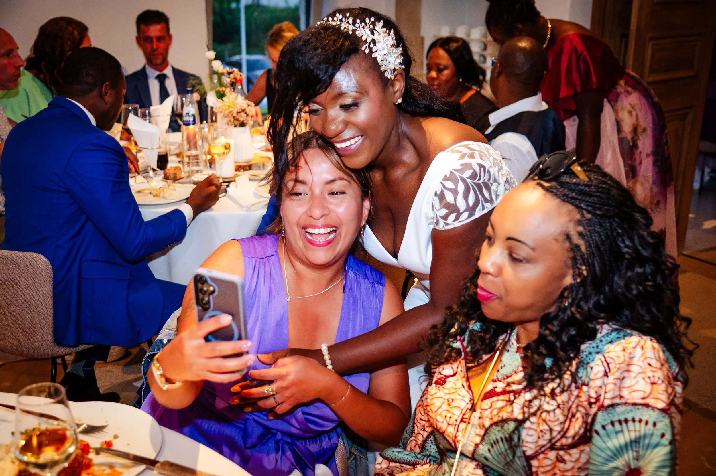 Vrouw in een witte jurk maakt een foto met haar telefoon terwijl ze lacht en twee andere vrouwen erbij zitten, op een feest met andere gasten aan tafel.
