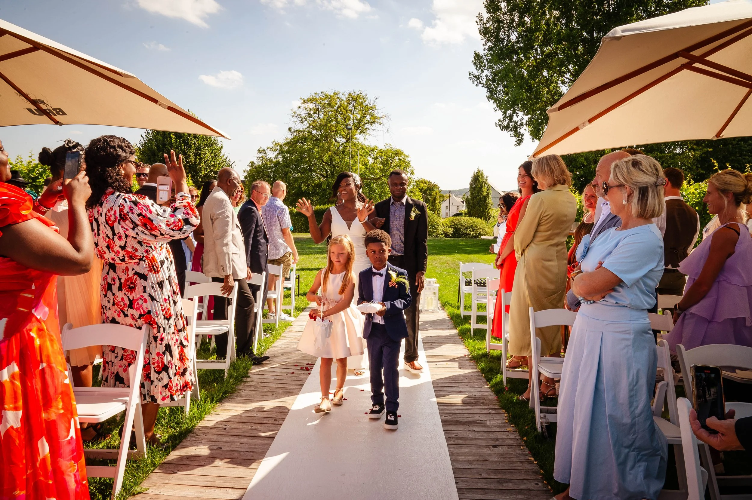 Omdat het een bruiloft is, lopen de bruidsmeisjes en de bruidegom langs de gasten. Het is een zonnige dag en de gasten staan en zitten onder grote parasols.