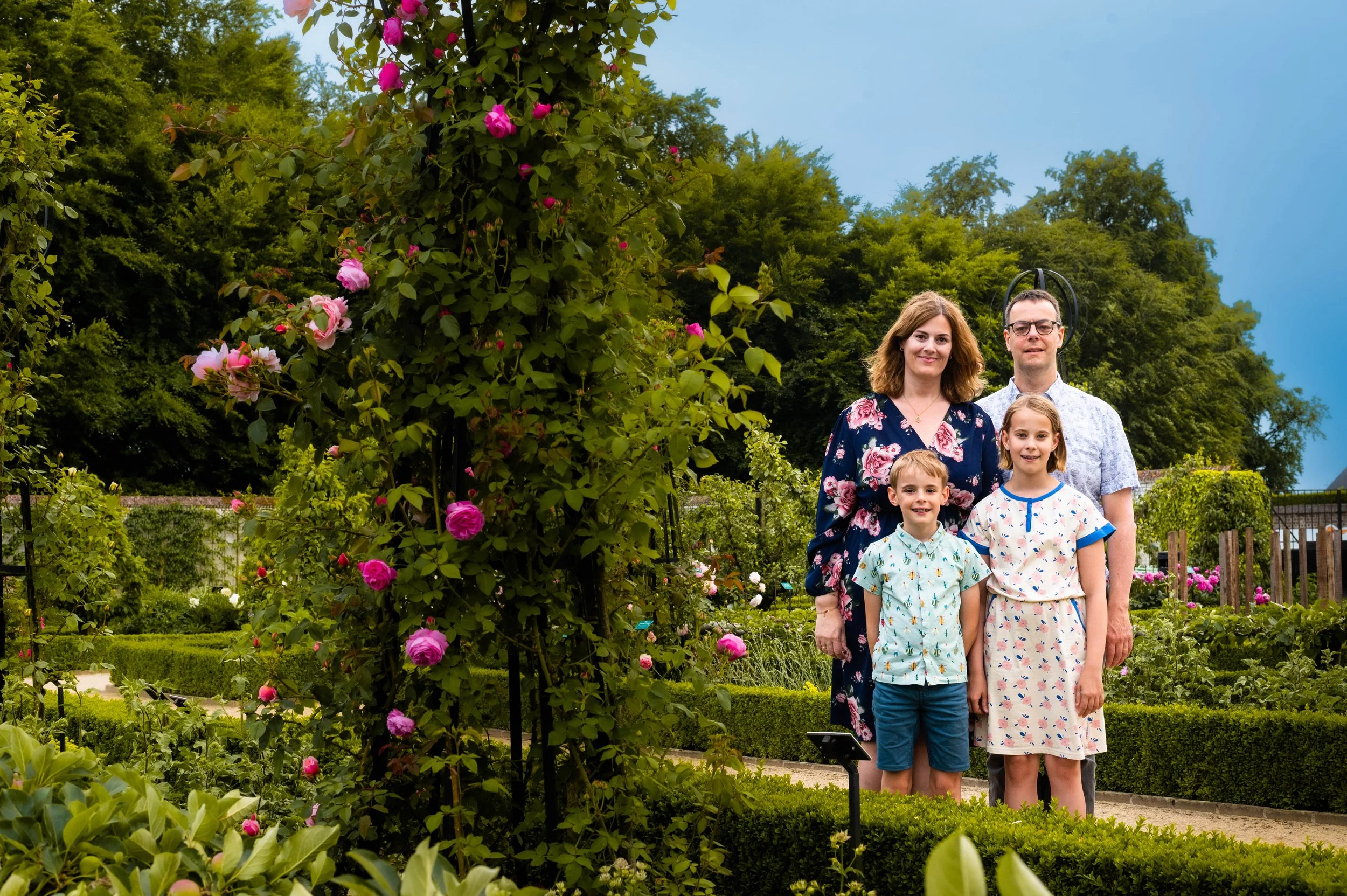 Familie van vier poseert in een tuin met rozen, omgeven door groene planten en struiken, onder een blauwe hemel.