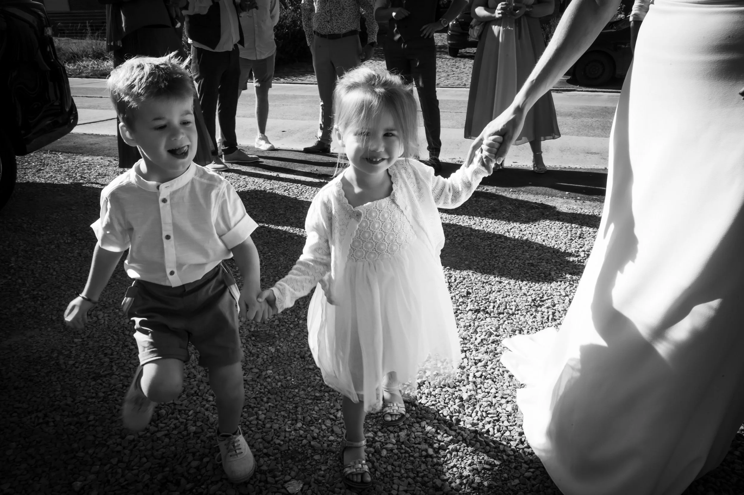 Twee jonge kinderen, een jongen en een meisje, maken plezier en lopen hand in hand met een volwassene tijdens een feest of bruiloft. De kinderen dragen nette kleding en lachen blij.