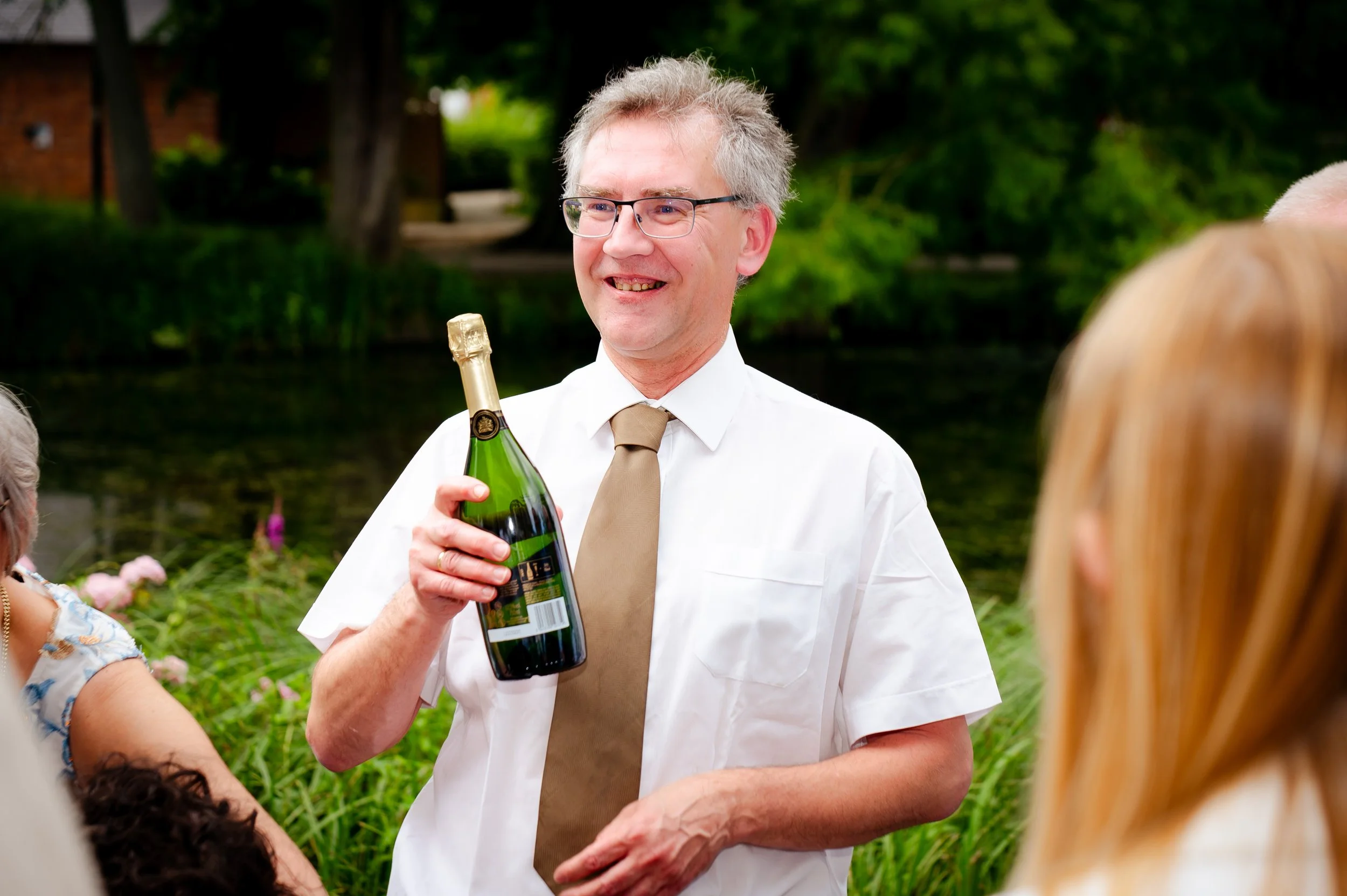 Man met bril en witte overhemd, lachend, houdt een fles champagne vast en spreekt met een groep mensen buiten nabij een meer, met groene bomen op de achtergrond.