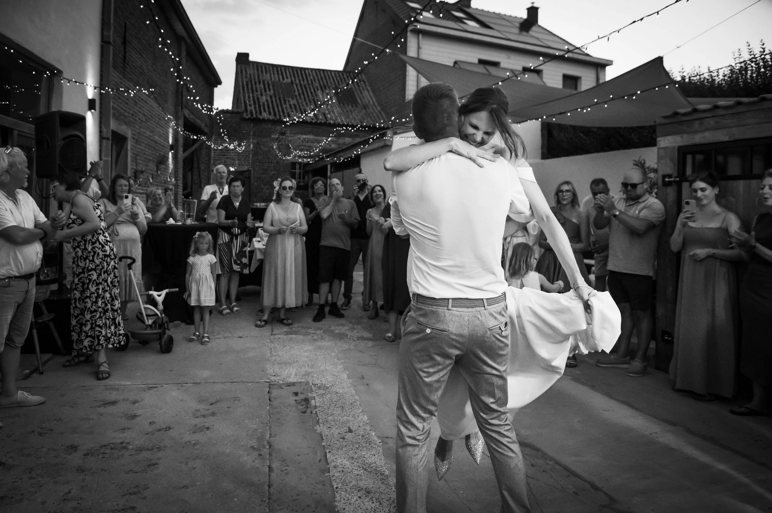 Een stel danst tijdens een bruiloft, omringd door vrienden en familie in een buitenruimte met decoraties en verlichting.
