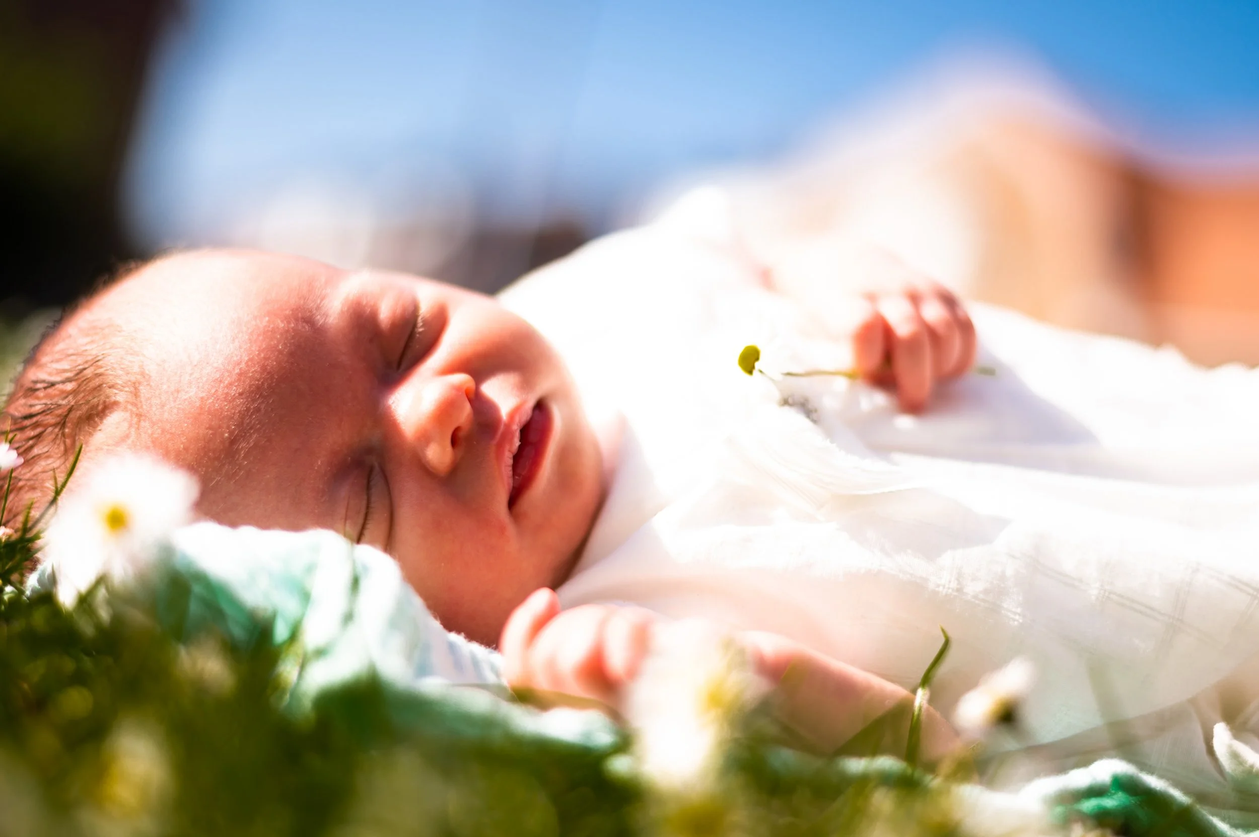 Close-up van slapende baby die in het gras ligt met bloemen, onder een blauwe lucht.