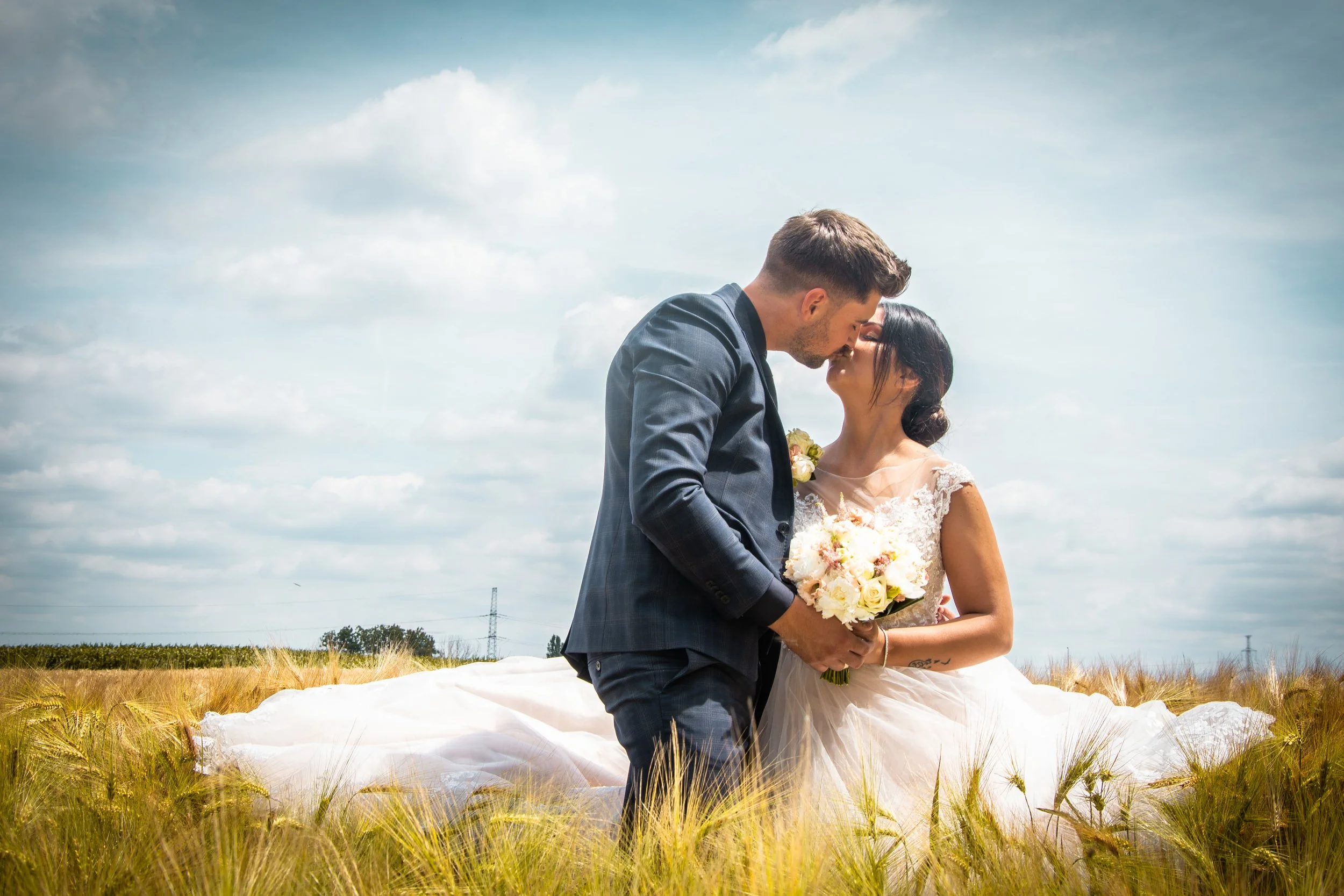 Koppel in huwelijkskleding die kussen in een veld met tarwe, de vrouw houdt een boeket bloemen vast.