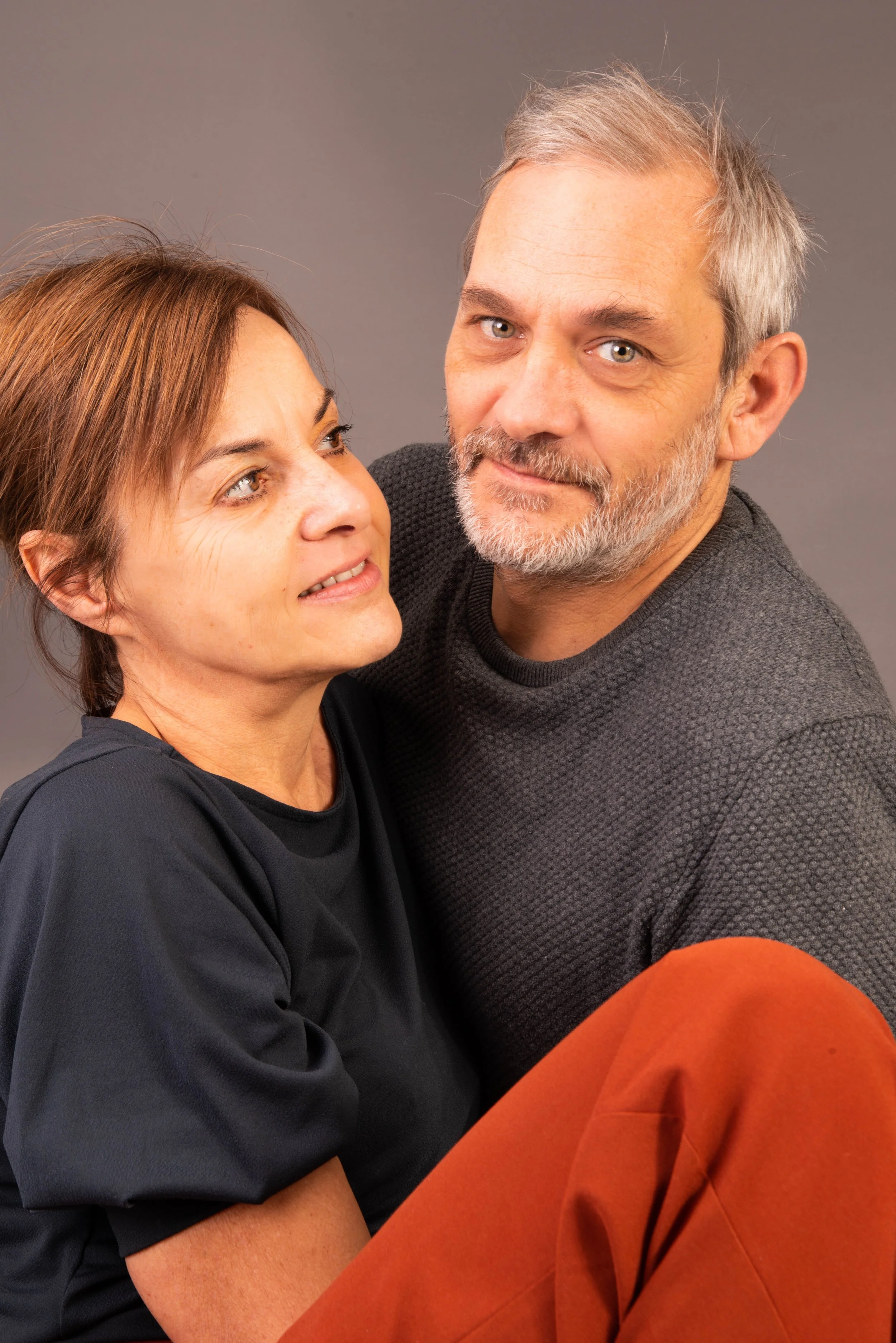 Een man en een vrouw kijken elkaar liefdevol aan. De vrouw heeft bruin haar en draagt een zwart shirt, de man heeft grijs haar en een grijze trui. Ze zitten dicht naast elkaar, tegen een effen grijze achtergrond.