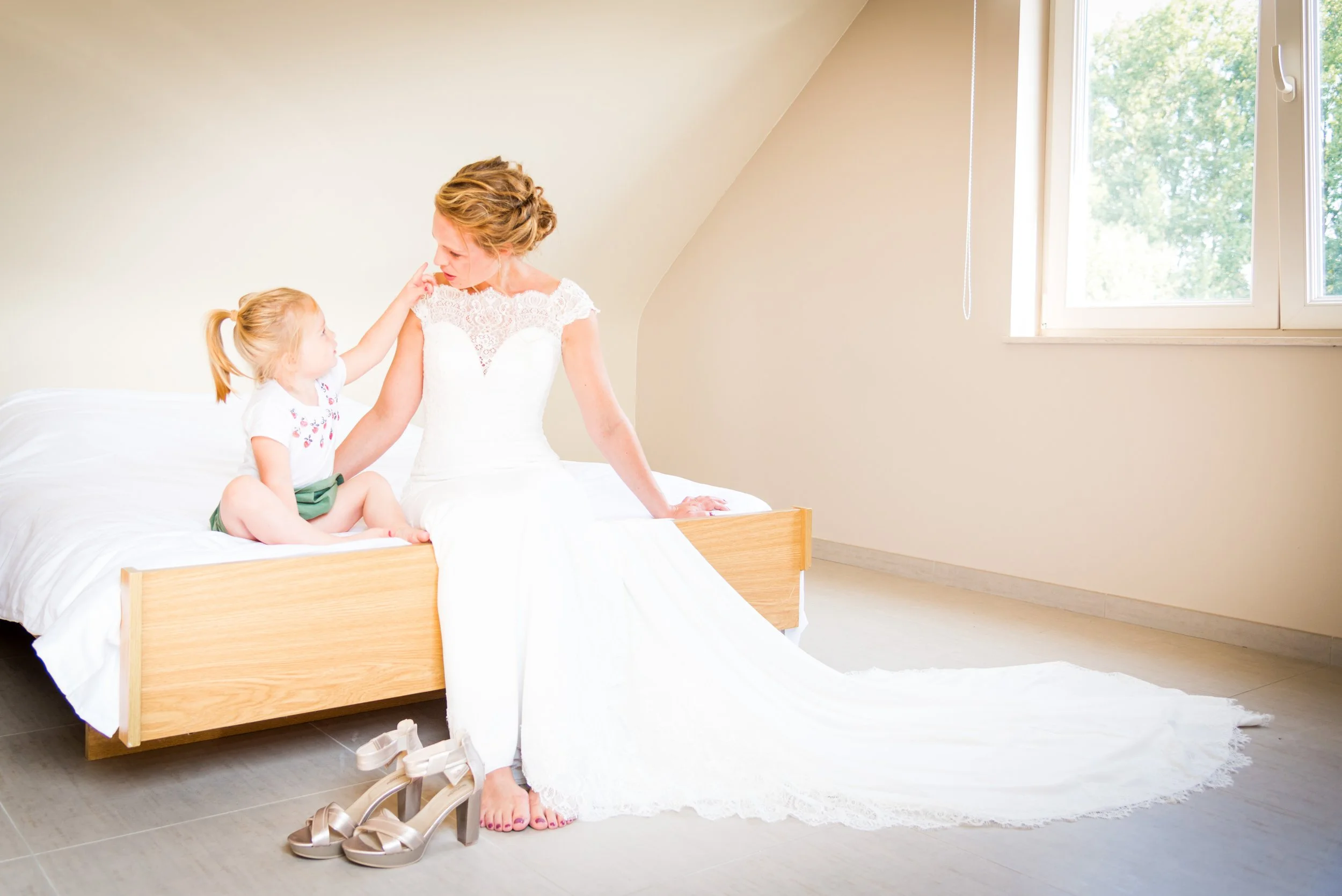 Vrouw in witte trouwjurk en een jonge meisje zitten op een bed, het meisje raakt aan de schouder van de vrouw. De schoenen van de vrouw liggen naast het bed.