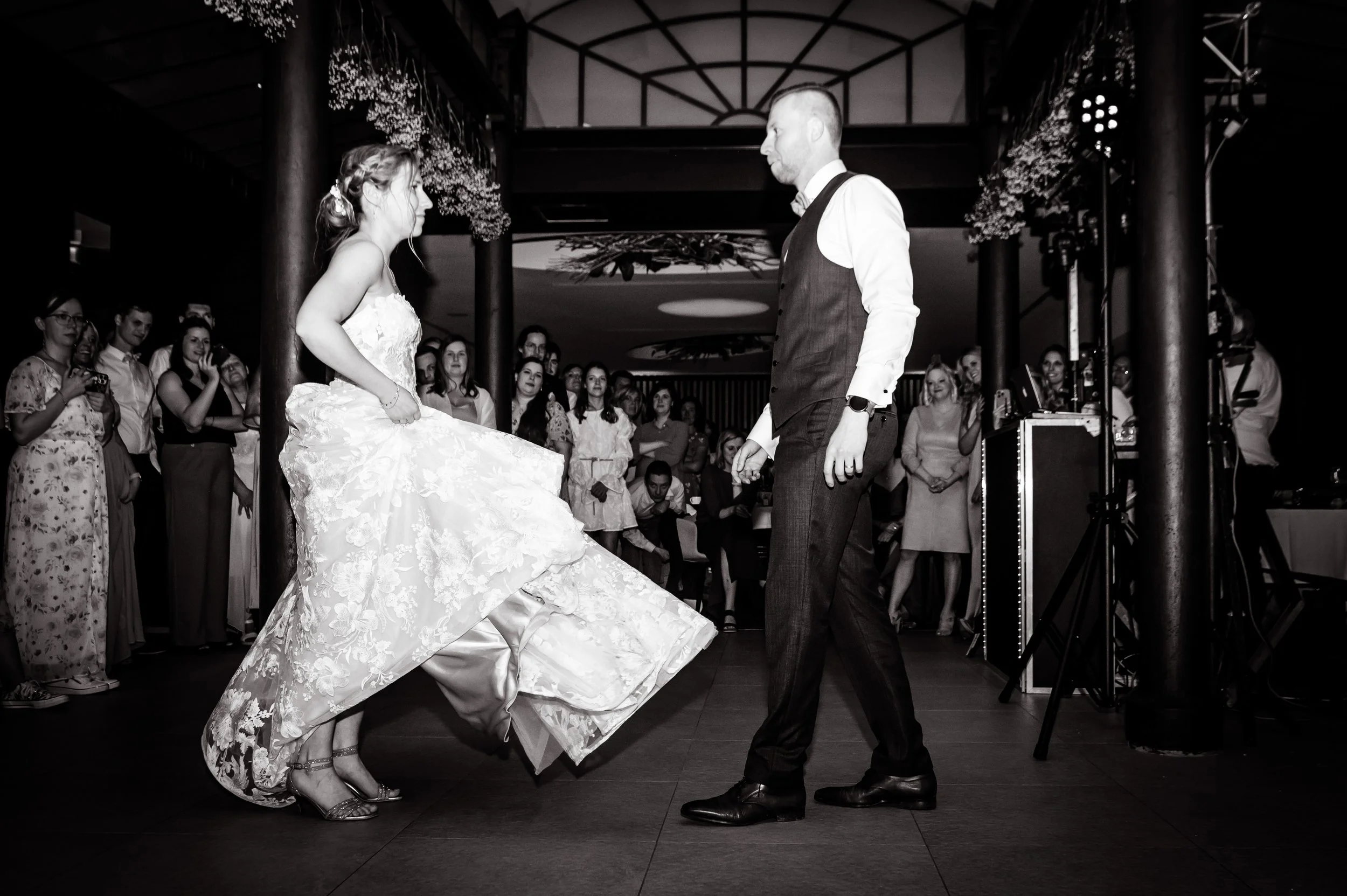 Een bruid en bruidegom dansen op de dansvloer tijdens een bruiloft, omringd door gasten die toekijken. Het is een zwart-wit foto.