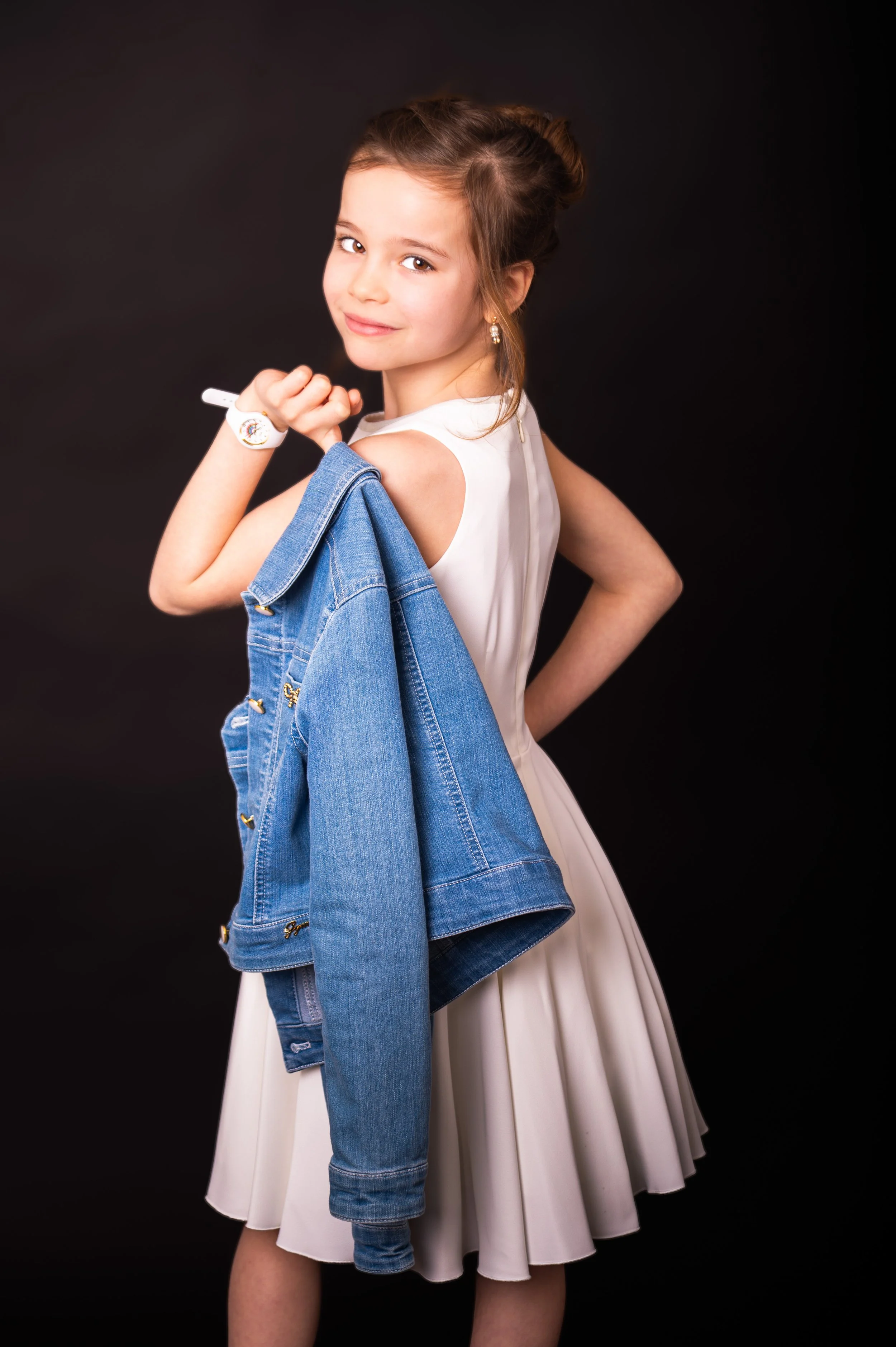 Jonge meisje met een witte jurk en een denim jas over haar schouder, poseert tegen een zwarte achtergrond.