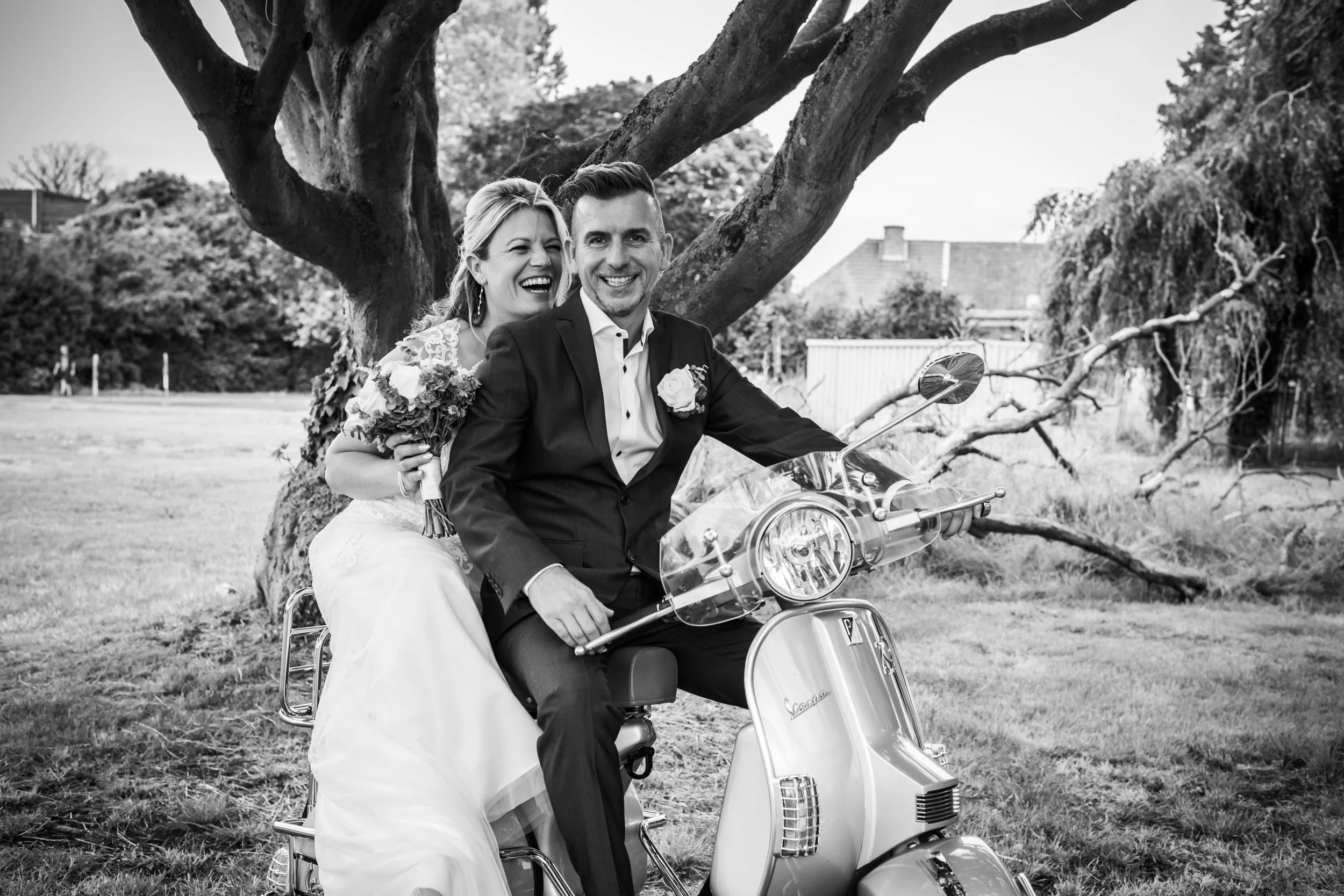 Een bruid en bruidegom die op een scooter zitten, onder een groot boom, beiden lachend, zwart-wit foto, buitenopnamen.