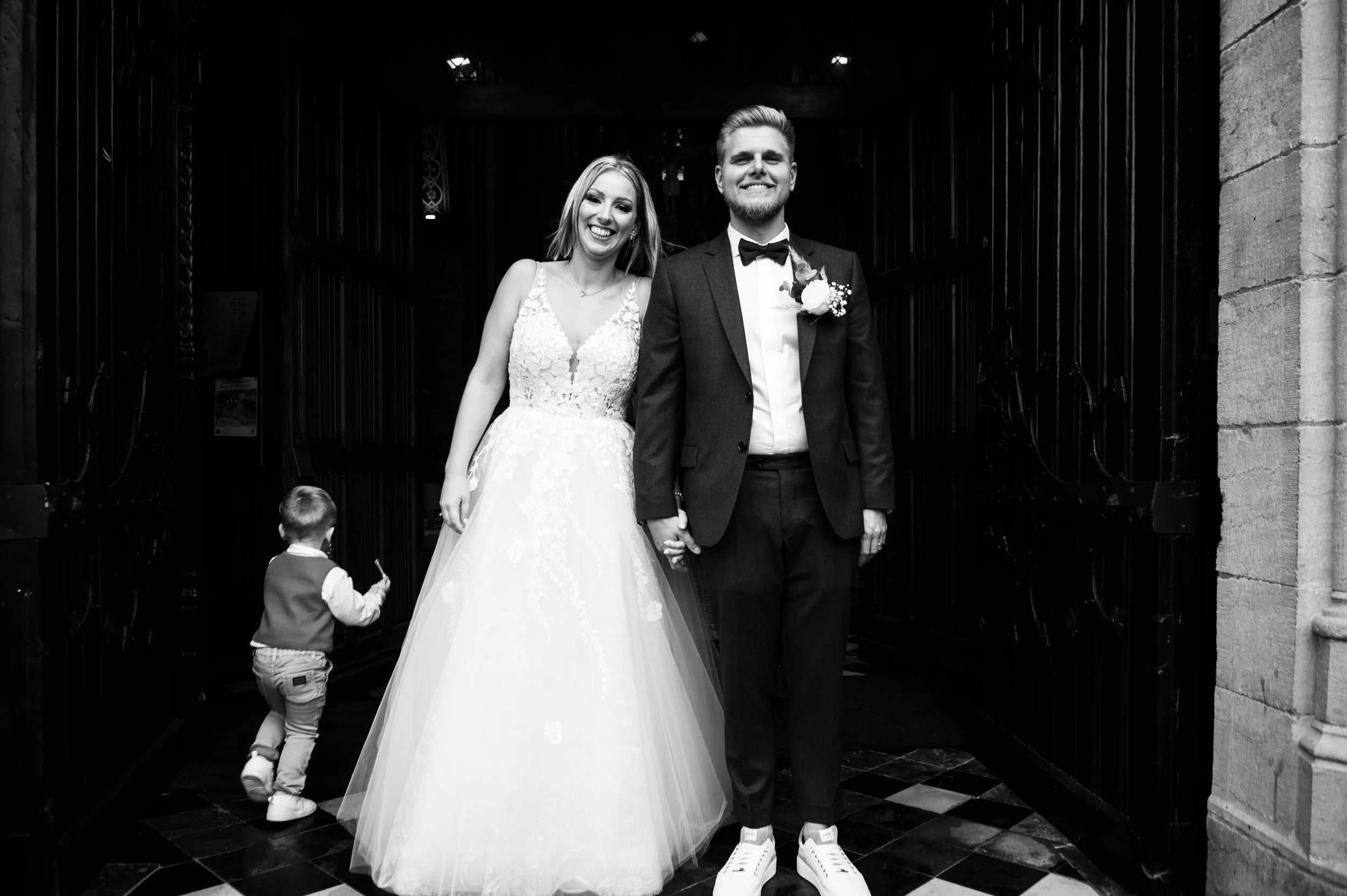 Bruidegom en bruid hand in hand poseren in een kerk, terwijl een kind op de achtergrond wegloopt.