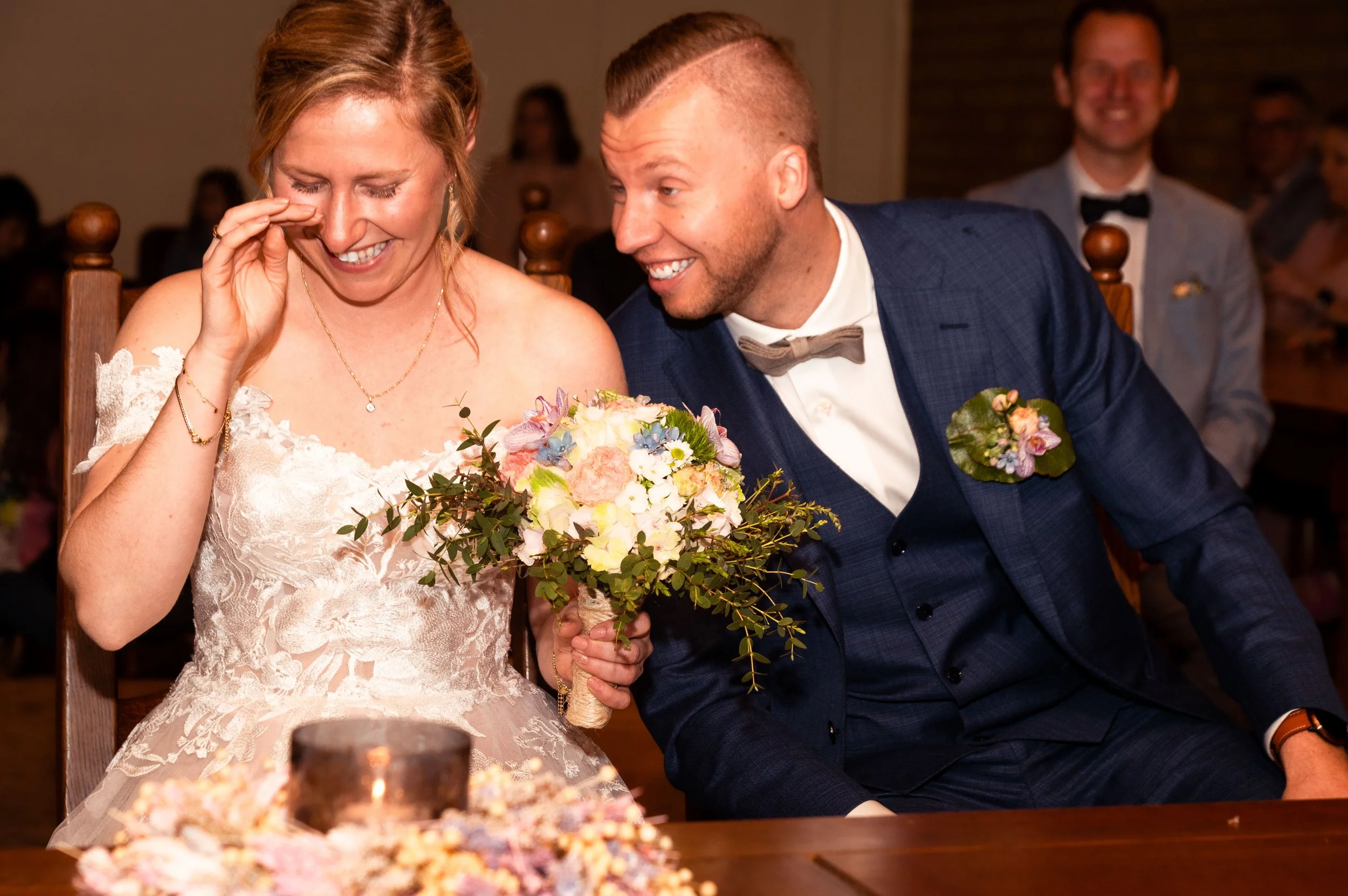 Een getrouwde vrouw en man lachen samen terwijl ze naar een bruidssboeket kijken tijdens hun bruiloft, omgeven door gasten.