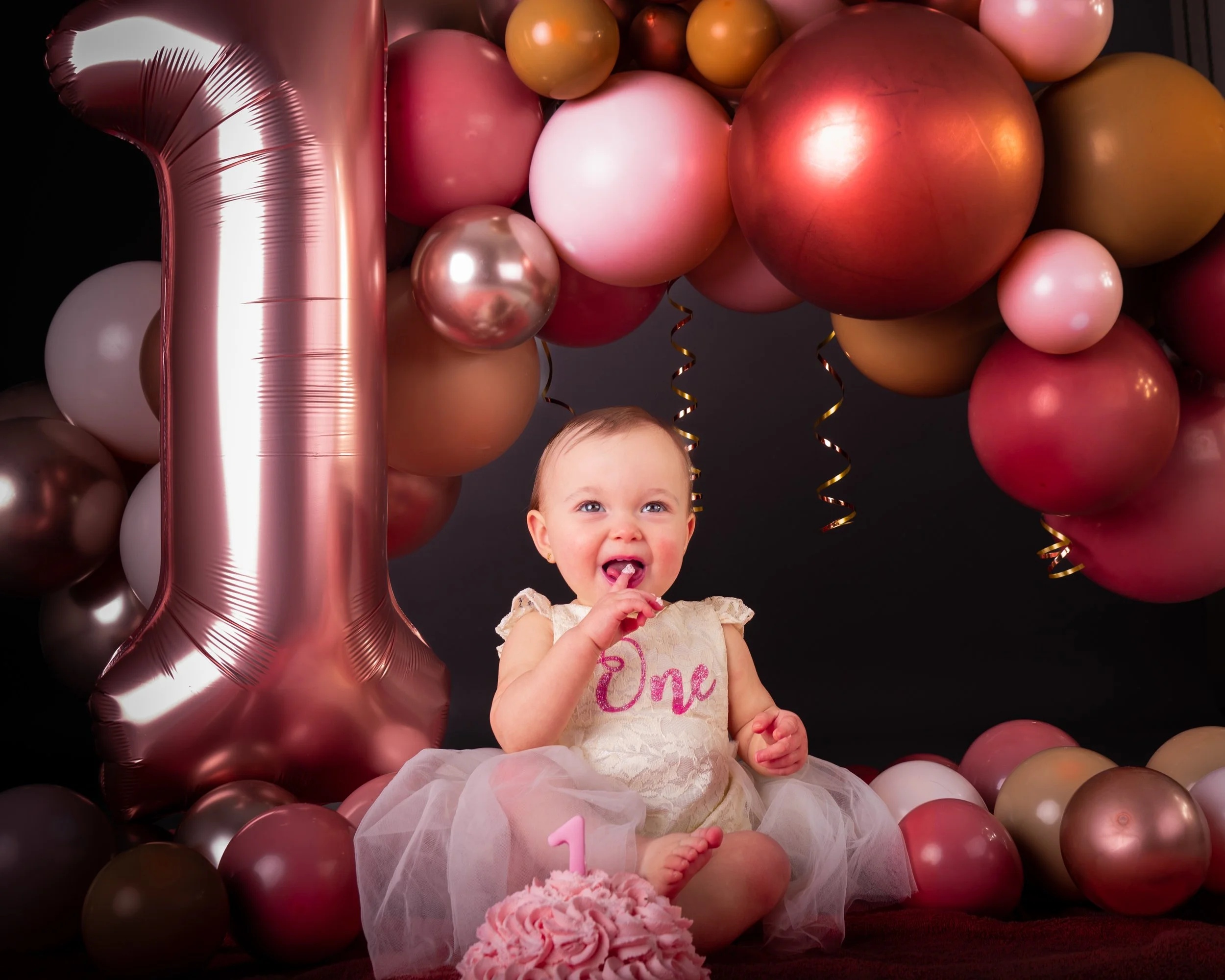 Een jonge meisje zit op de grond tijdens haar eerste verjaardag, omringd door roze, gouden en metallic ballonnen en een grote balon in de vorm van de cijfer 1, met een taart met een geboortecijfer 1 en een 'One' tekst op haar jurk.