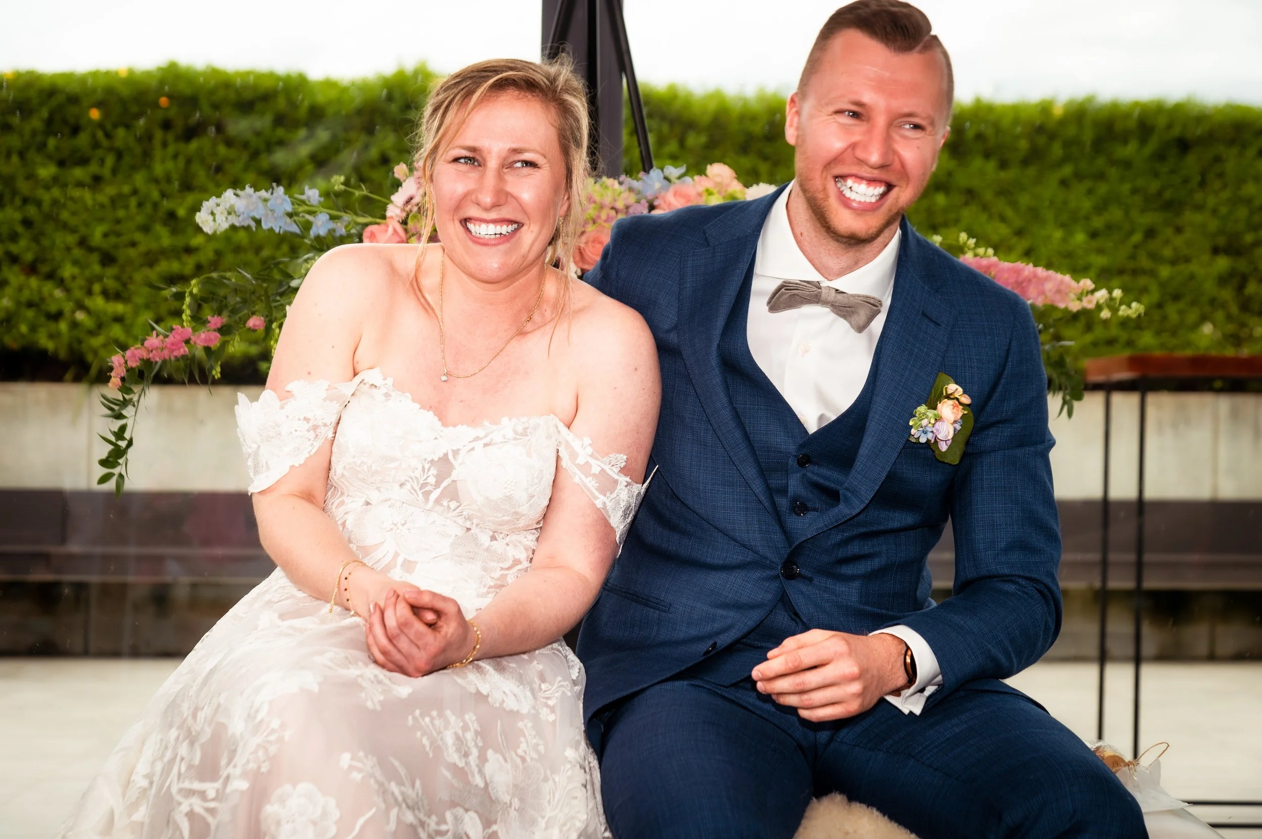 Een bruid en bruidegom zitten samen op een bank, lachend. Zij draagt een wit kanten bruidsjurk en hij een blauwe pak met een grijze strik en bloemen als boutonnière. Achtergrond met groen en bloemen.