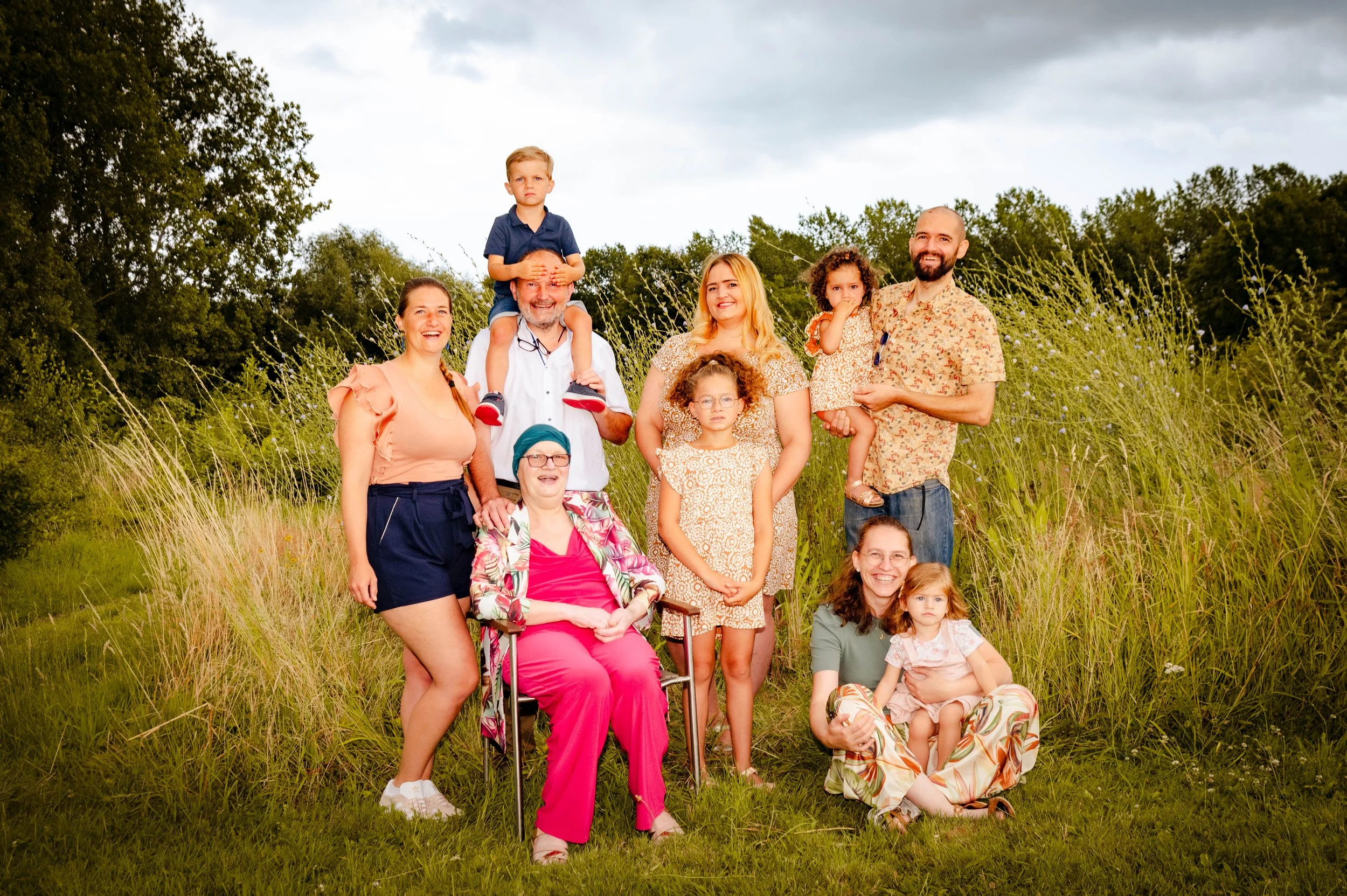 Groep familie die buiten in een veld poseert, met groene bomen en een bewolkte hemel op de achtergrond.