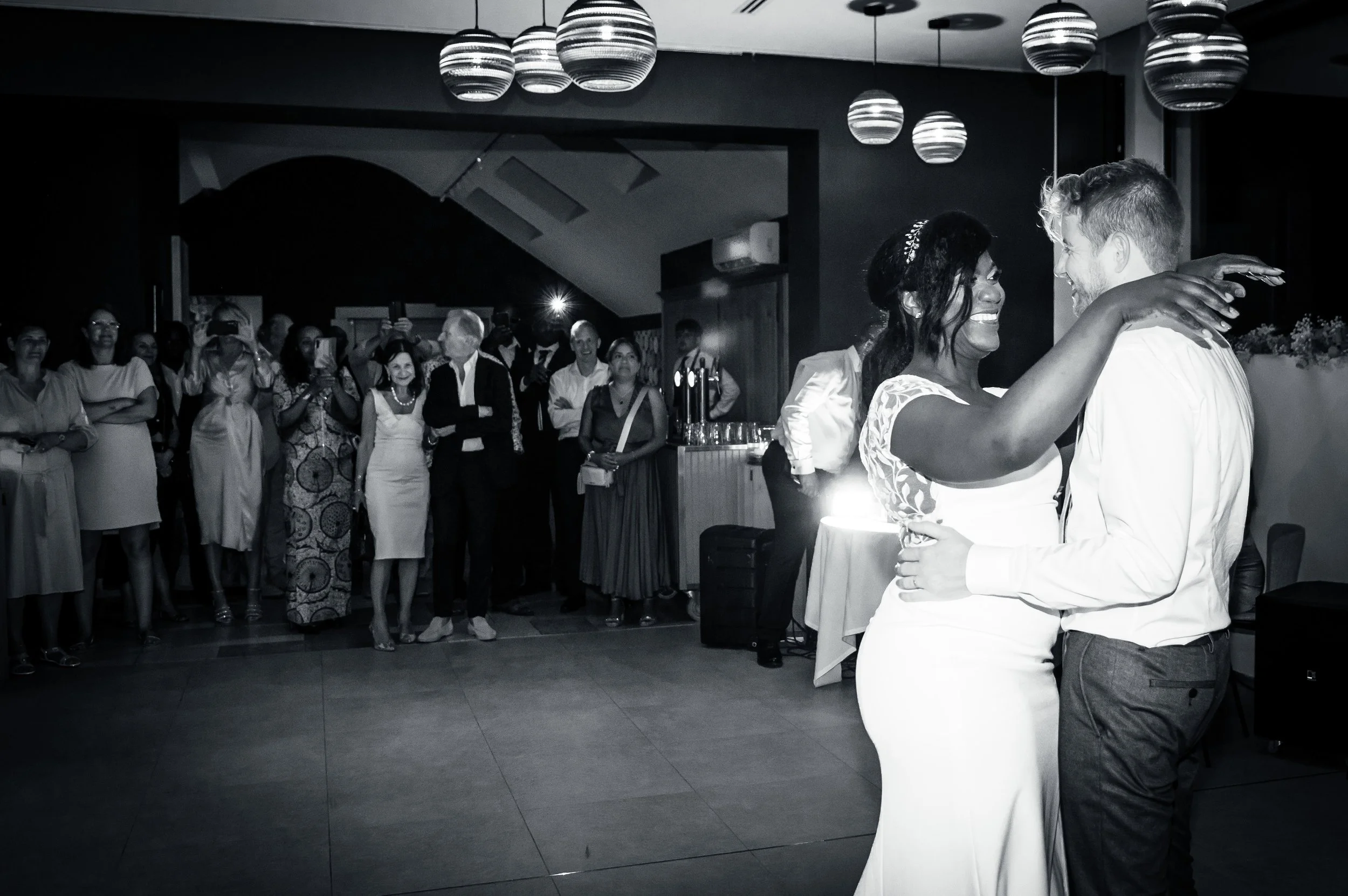 Een zwarte en witte foto van een bruid en bruidegom die dansen, omringd door gasten tijdens een bruiloft. De gasten kijken toe en nemen foto's.