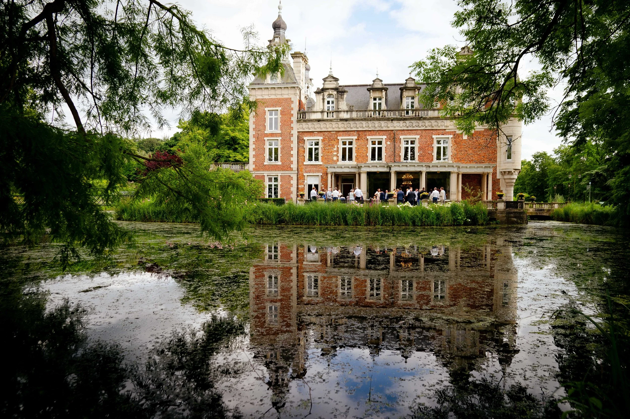 Een groot kasteel met een waterpartij op de voorgrond en een groep mensen die binnen in de patio staan, omgeven door groene bomen.
