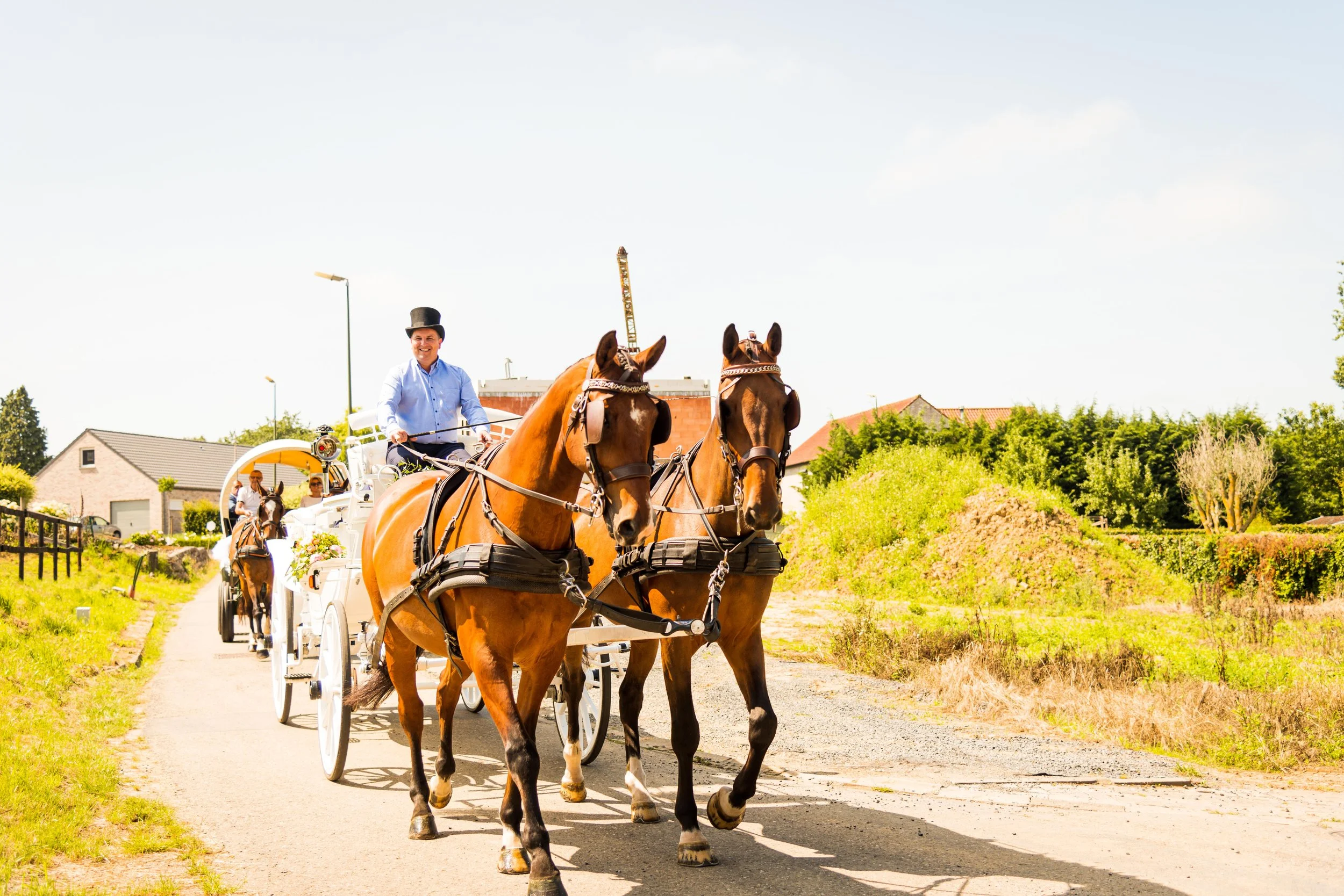 Mensen rijden in een koets met paarden door een landelijk gebied met groene velden en huizen onder een blauwe hemel.