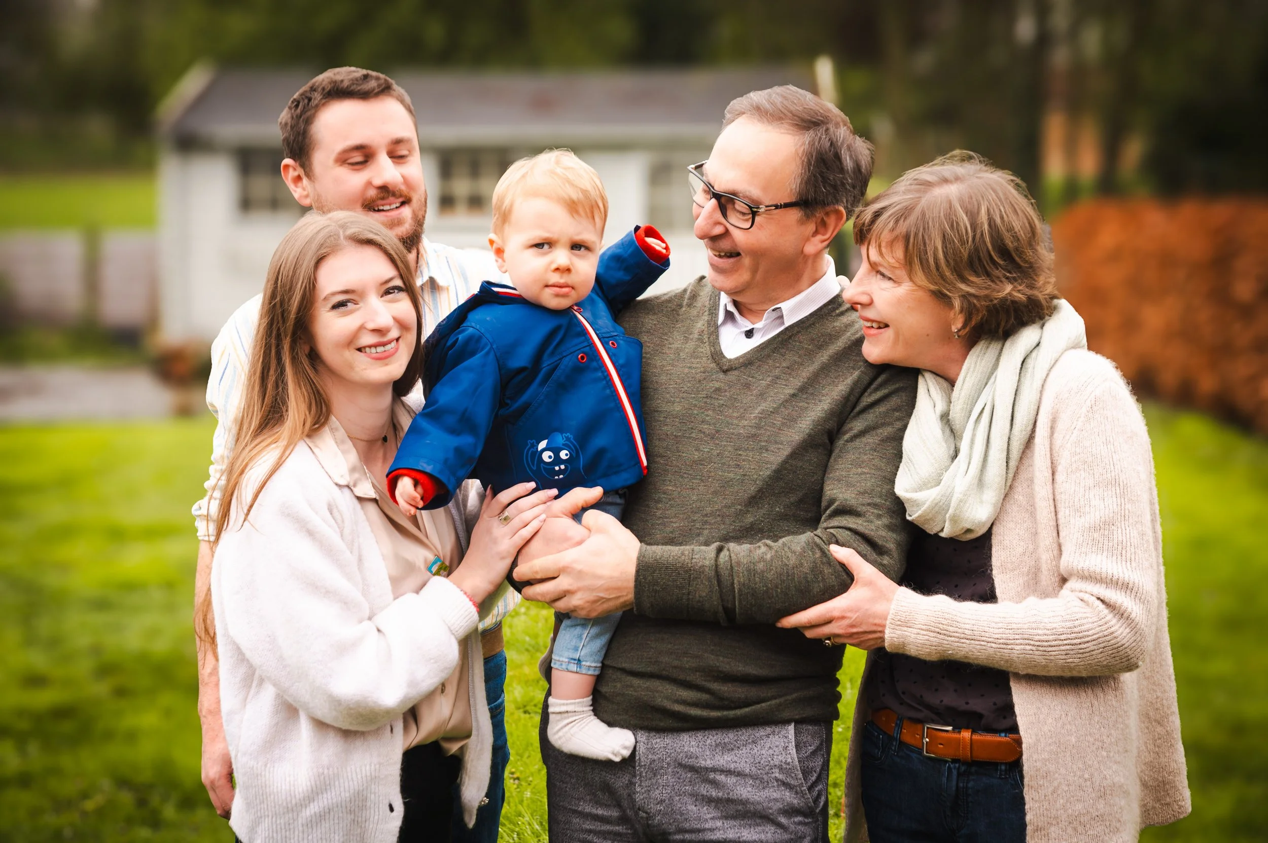 Een forward van een familie van vijf die buiten op het gras samen lachen en een jonge jongen vasthouden, met bomen en een huis op de achtergrond.