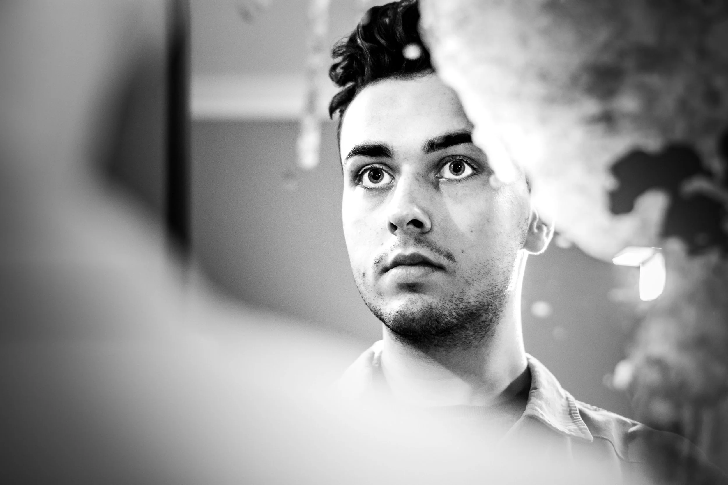 Zwart-wit foto van een jonge man met donkere haar, volop in close-up, die naar zichzelf in een spiegel kijkt.