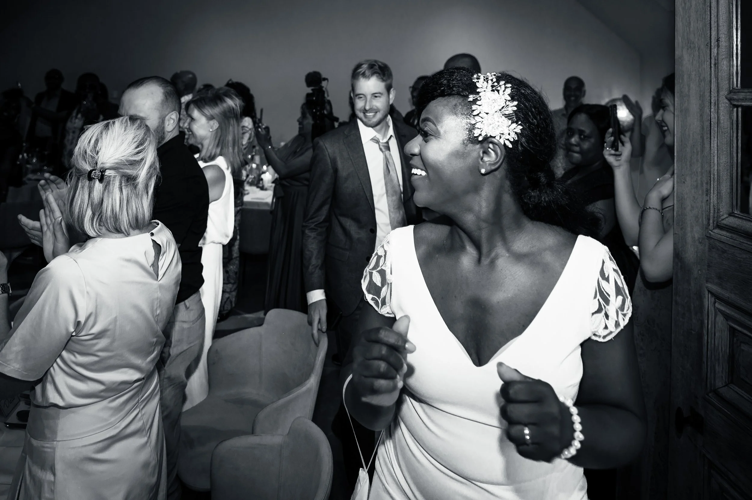 Feestvierende mensen op een feest, meeste mensen lachen en klappen, centrale vrouw in witte jurk met bloemen in haar haar die danst en glimlacht.