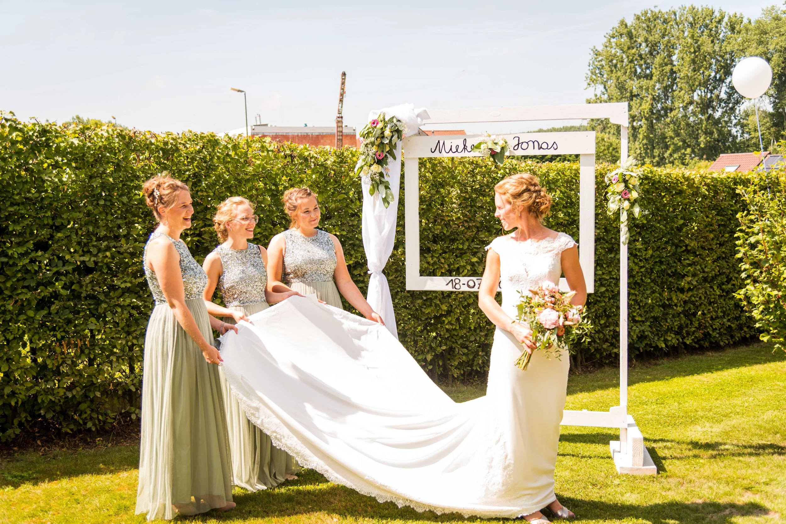 Een bruid met een witte jurk en een bloemenboeket krijgt een afscheidscadeau van drie vrouwen in een tuin met groene heg, onder een witte foto-achtergrond met bloemen, tijdens een zonnige dag.