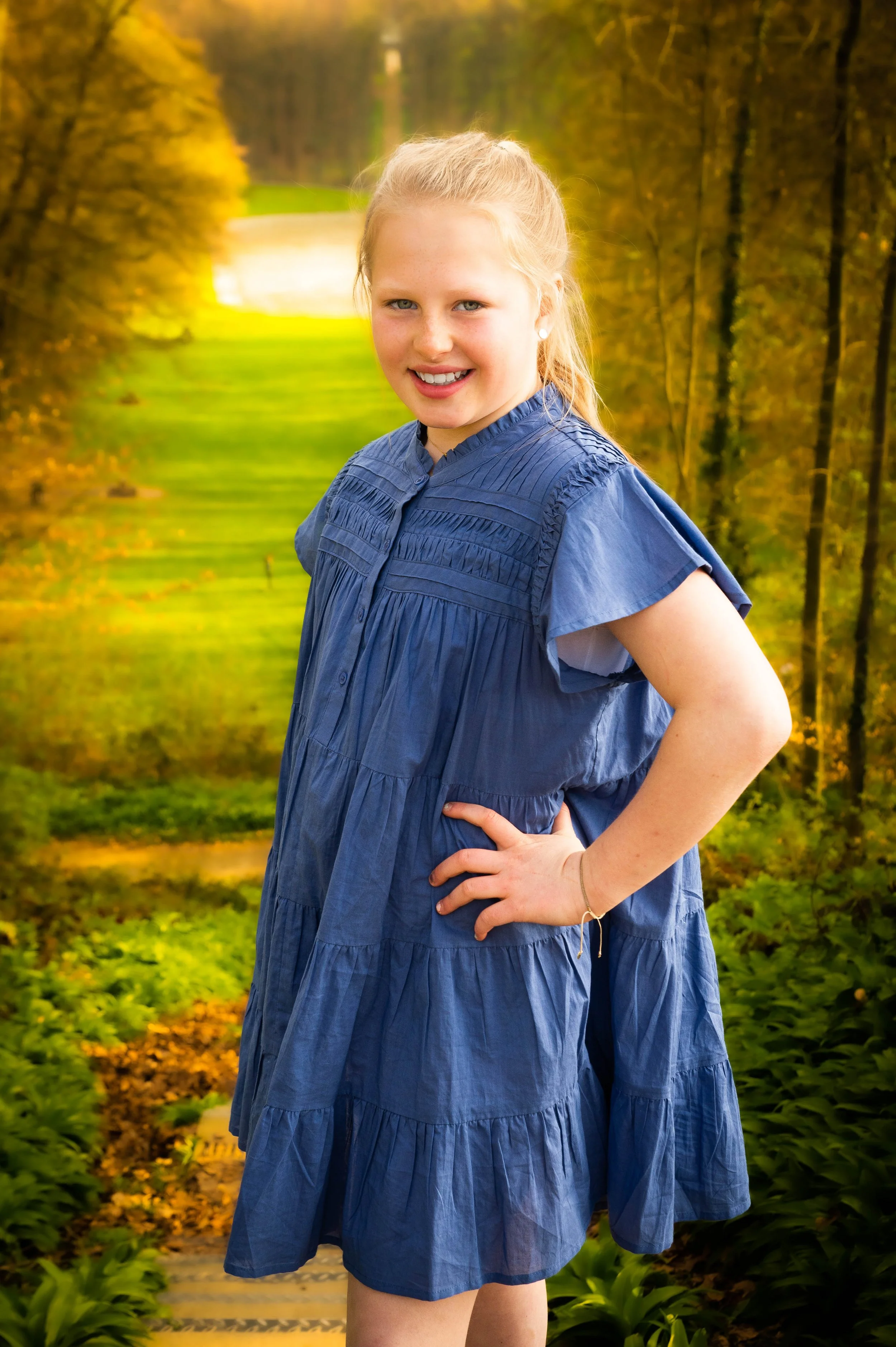Een lachende jonge vrouw met blond haar in een blauwe jurk die poseert voor een bosachtergrond.