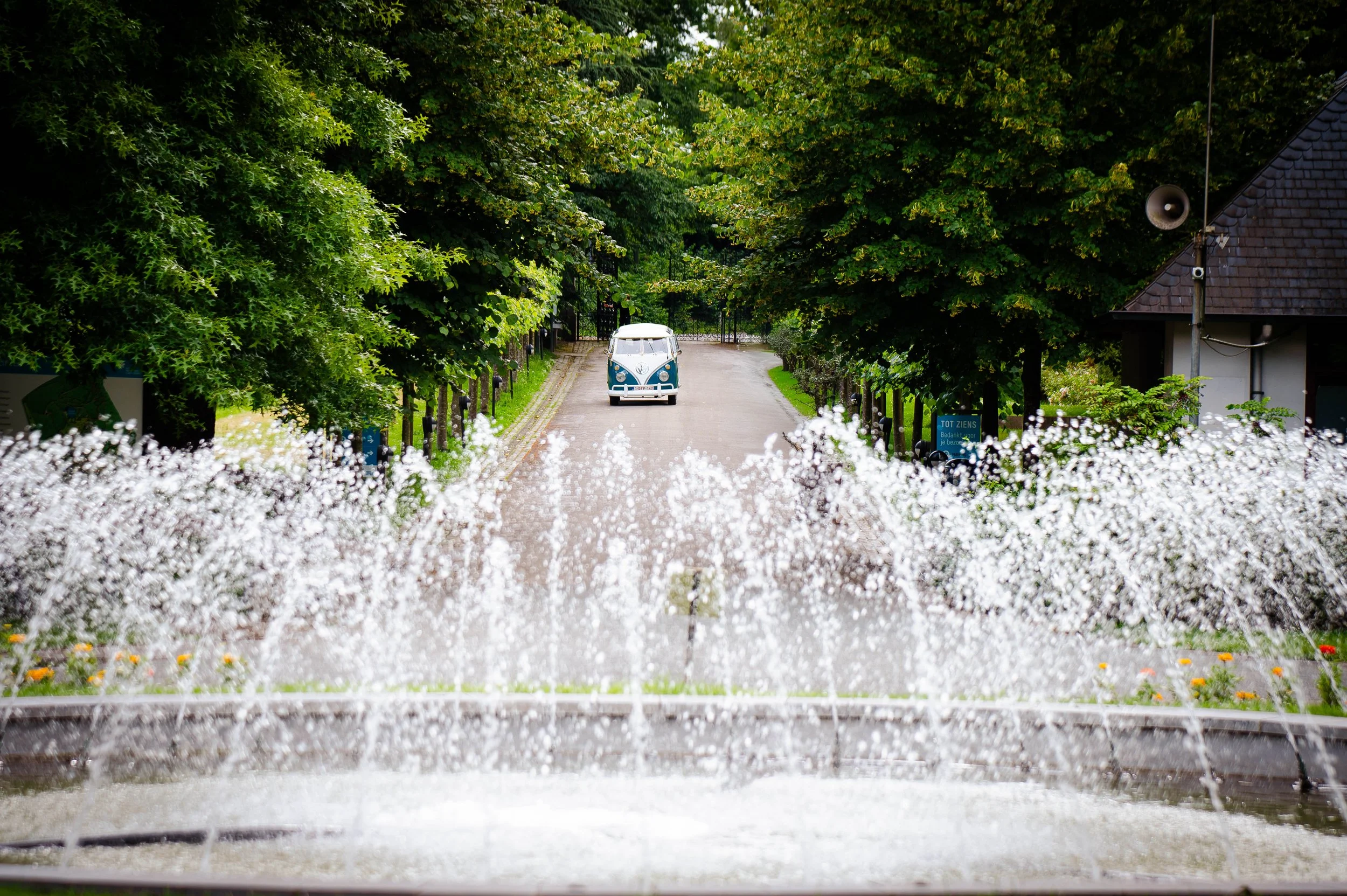 Een schoolplein met een blauwe Volkswagen bus, omgeven door groene bomen, zichtbaar achter een fontein met waterstralen.