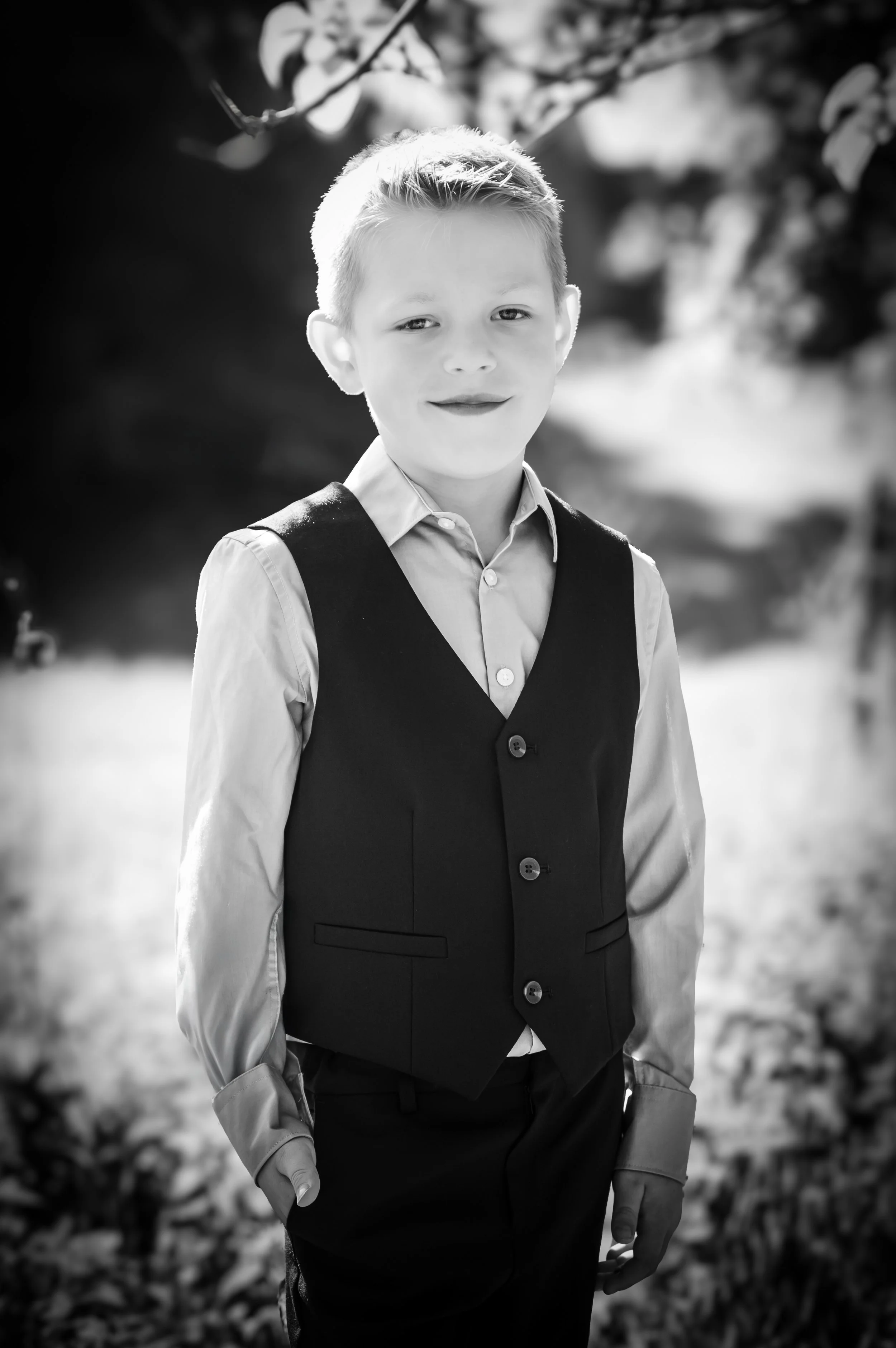 Portret van een jongen in formele kleding, staand buiten met een donkere achtergrond van bladeren.